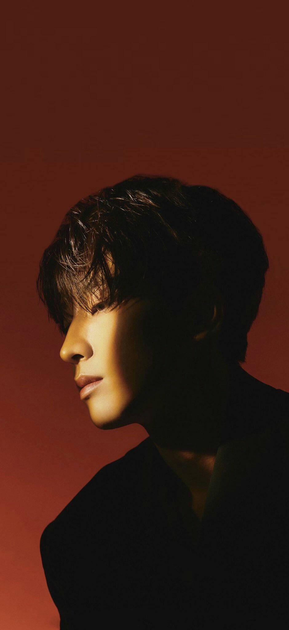 Wonwoo 桌布 壁紙 — 一名年輕男子的側面，沐浴在溫暖光線中，背景為深紅色，帶有戲劇性的陰影。