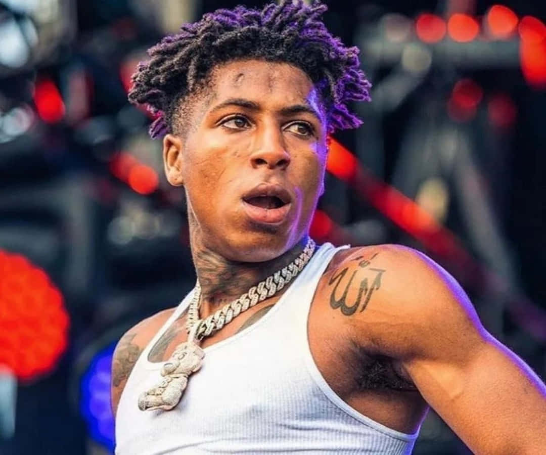 NBA YoungBoy 桌布 壁紙 — 一名年輕男子的特寫，他有紫色染髮的辮子，身穿白色背心和粗鏈項鍊，背景是模糊的舞台和紅燈。