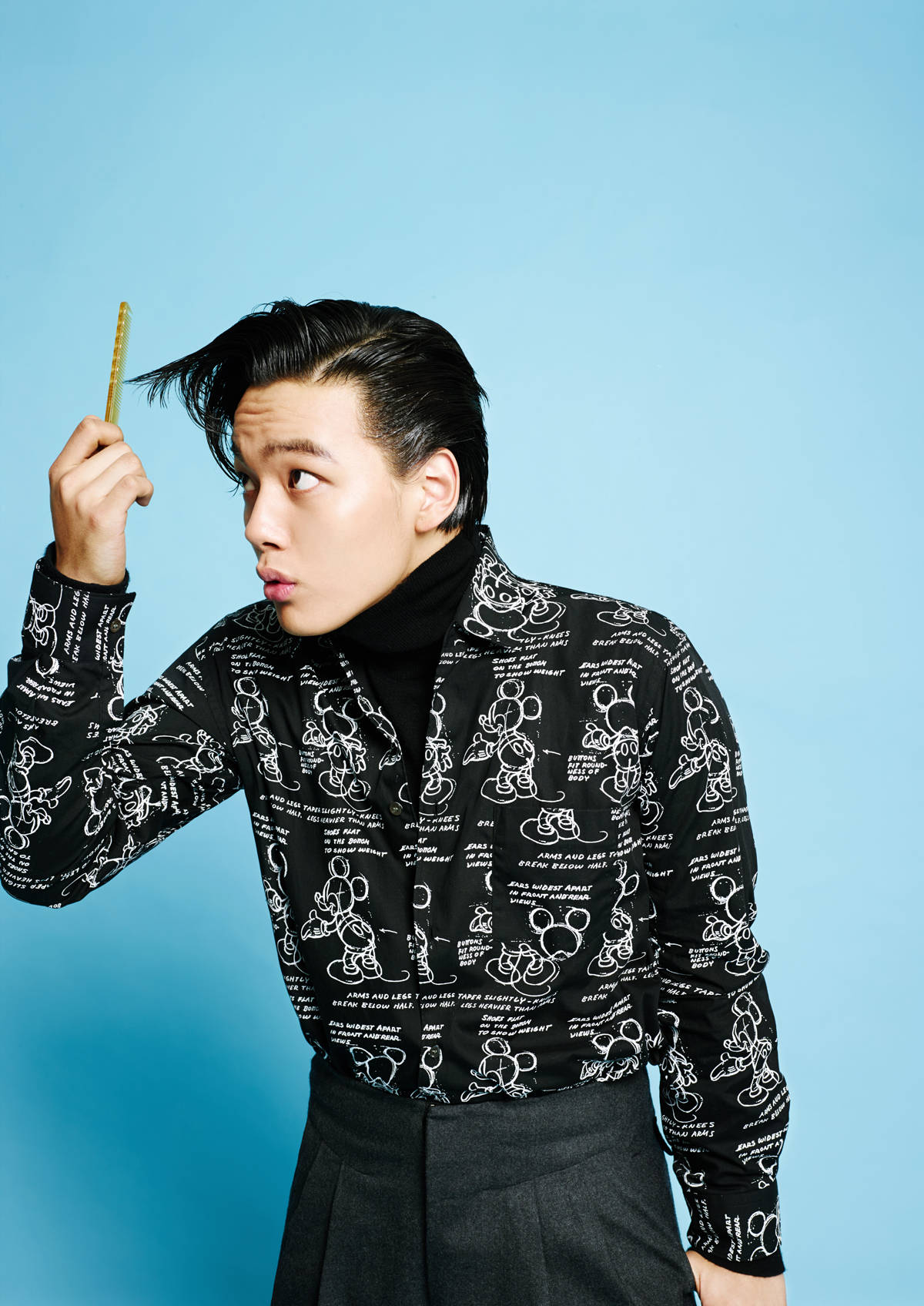 Jin 桌布 壁紙 — 一名年輕男子穿著印有米奇老鼠圖案的黑色襯衫，在純藍色背景前，頑皮地拿著梳子靠近他造型過的頭髮。