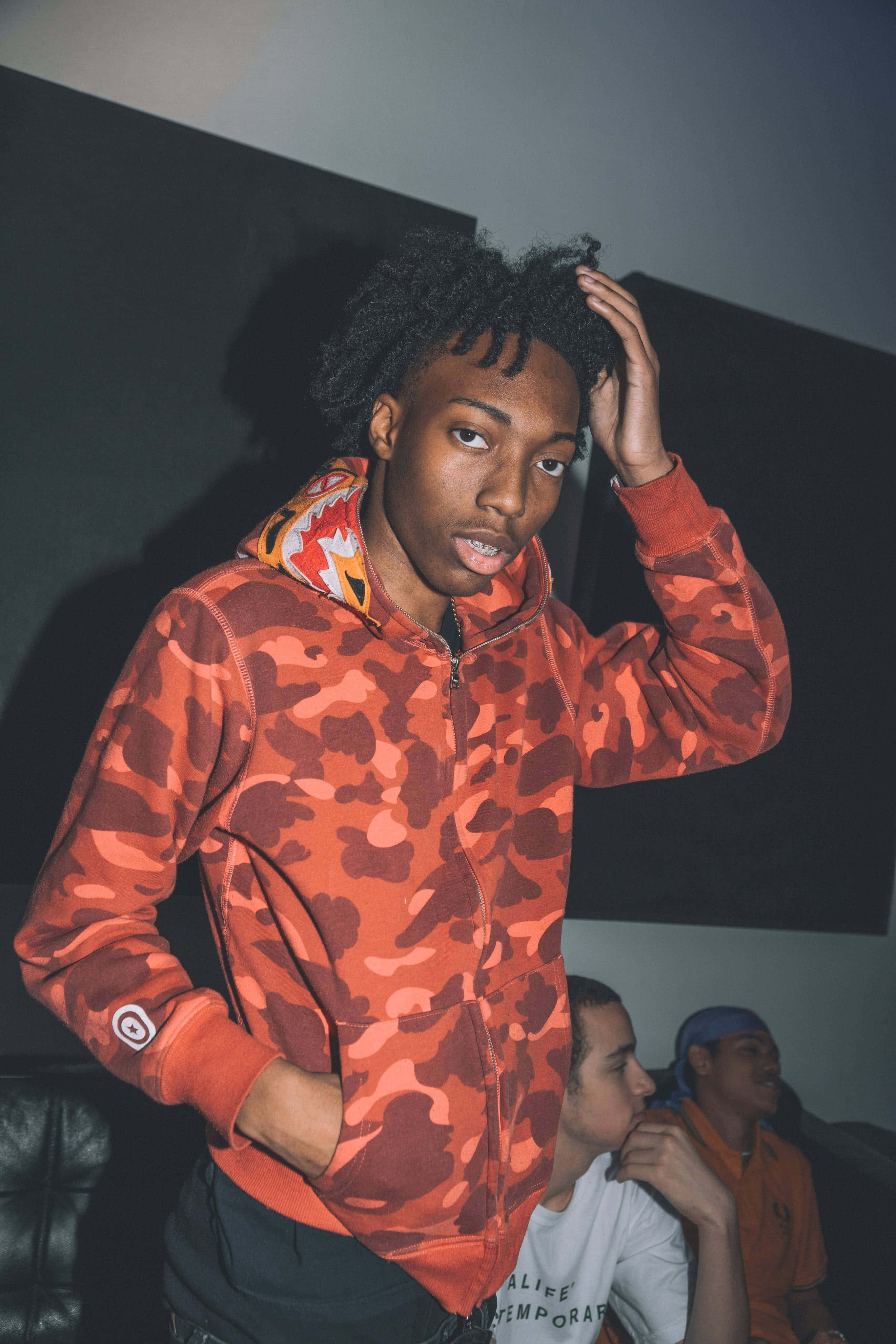 Rappers 桌布 壁紙 — 一名年輕男子穿著大膽的橙色迷彩連帽衫，在戲劇性的燈光下自信地在室內擺姿勢，一手放在頭髮上。