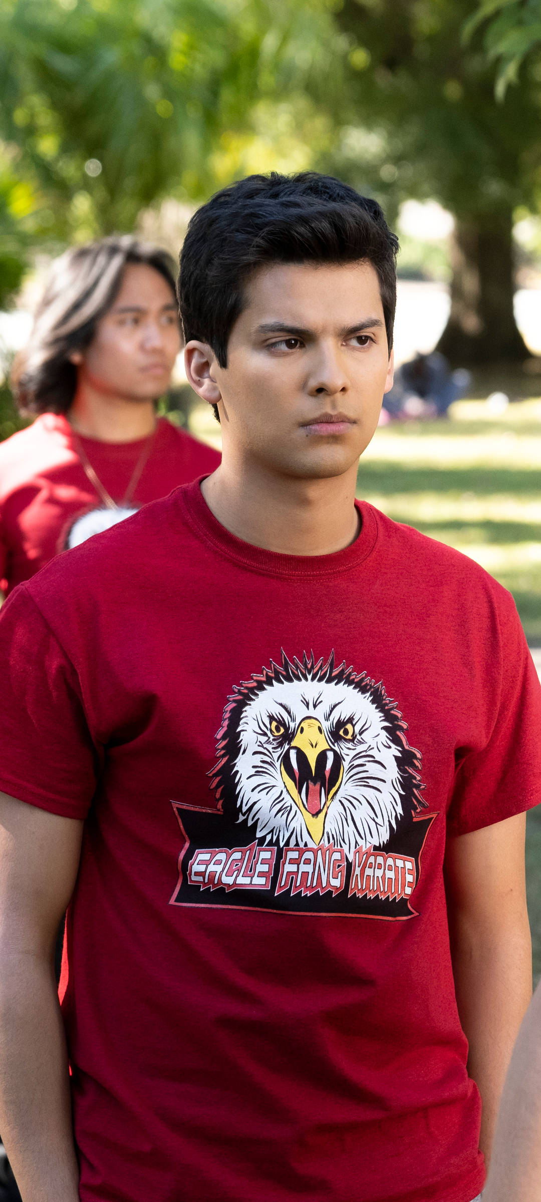 Eagle Logo 桌布 壁紙 — 一名年輕男子穿著紅色Eagle Fang Karate T恤，表情嚴肅地站在戶外，背景中另一個人模糊不清。