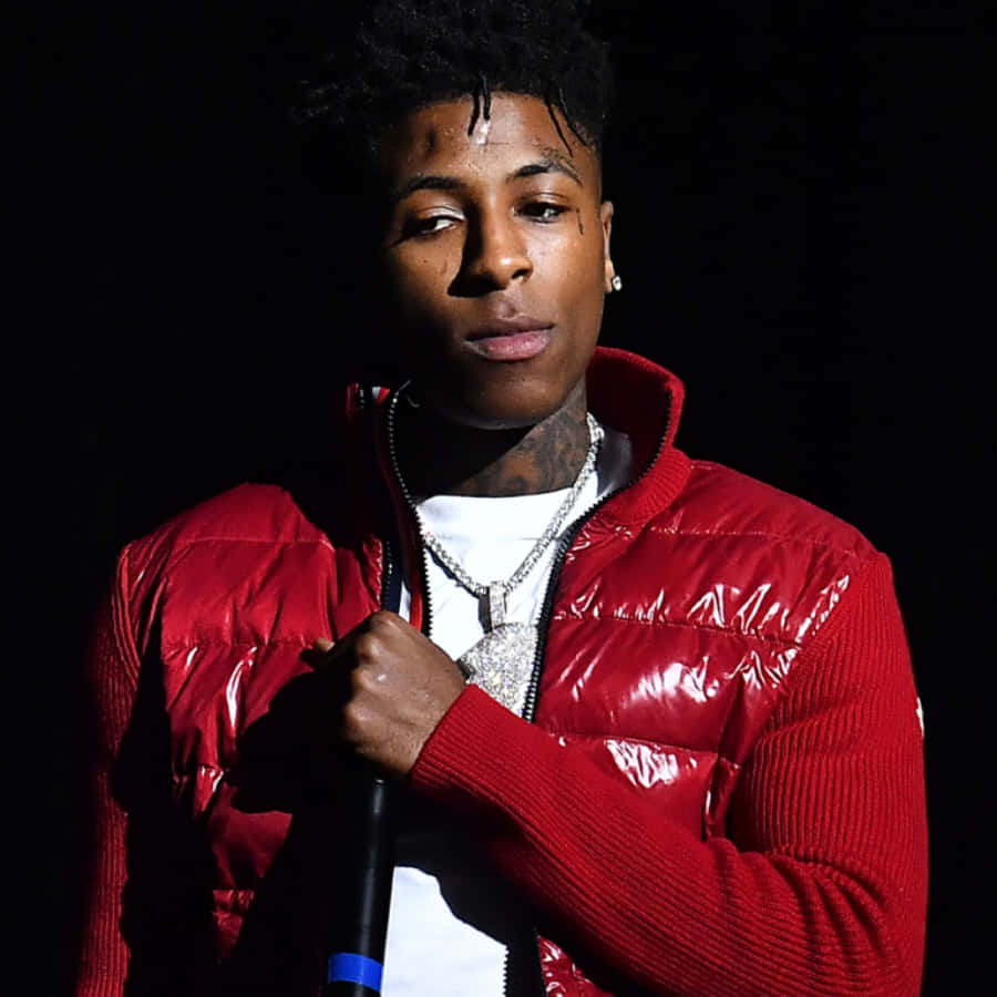 NBA YoungBoy 桌布 壁紙 — 一名年輕男子穿著閃亮的紅色羽絨外套，在戲劇性的舞台燈光下，對著黑色背景握著麥克風。