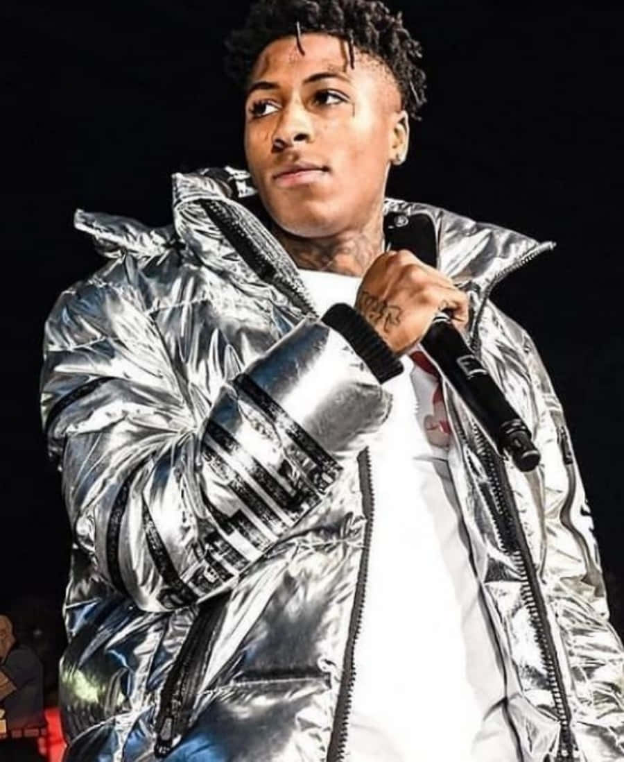 NBA YoungBoy 桌布 壁紙 — 一名年輕男子穿著閃亮的銀色羽絨外套，在黑暗背景下拿著麥克風。