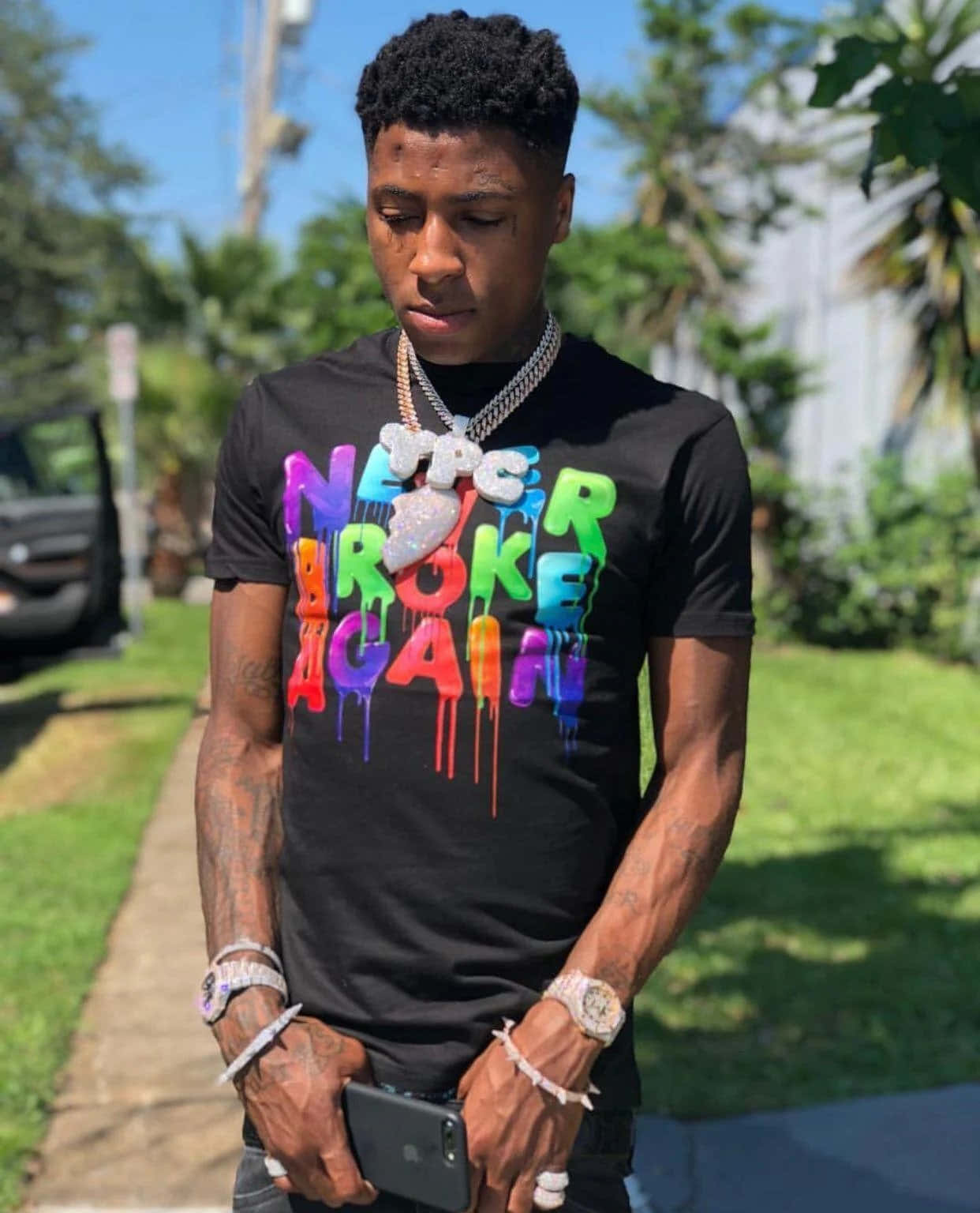 NBA YoungBoy 桌布 壁紙 — 一名年輕男子穿著黑色T恤，上面有彩色滴落文字，在陽光明媚的日子裡站在戶外拿著電話。
