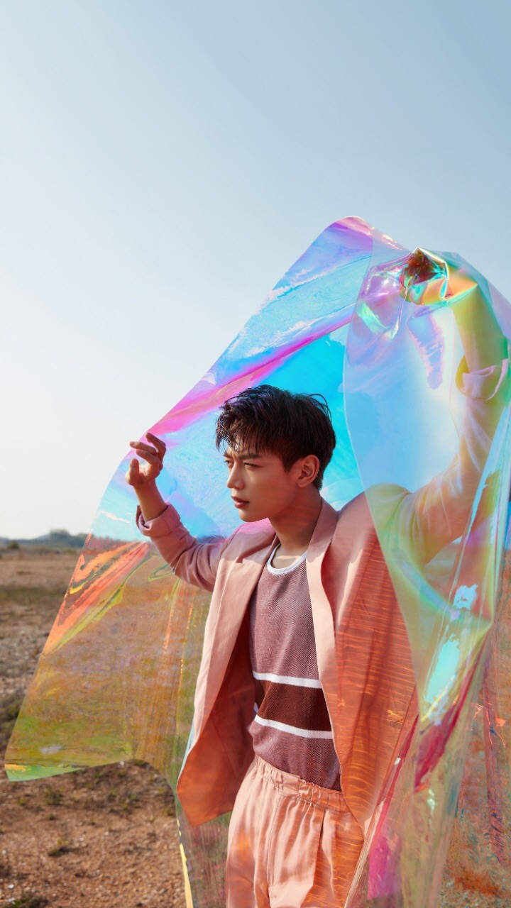Shinee 桌布 壁紙 — 一名年輕男子站在乾燥的田野中，手持一塊巨大的彩虹色布料，在清澈的藍天下閃爍著虹彩。