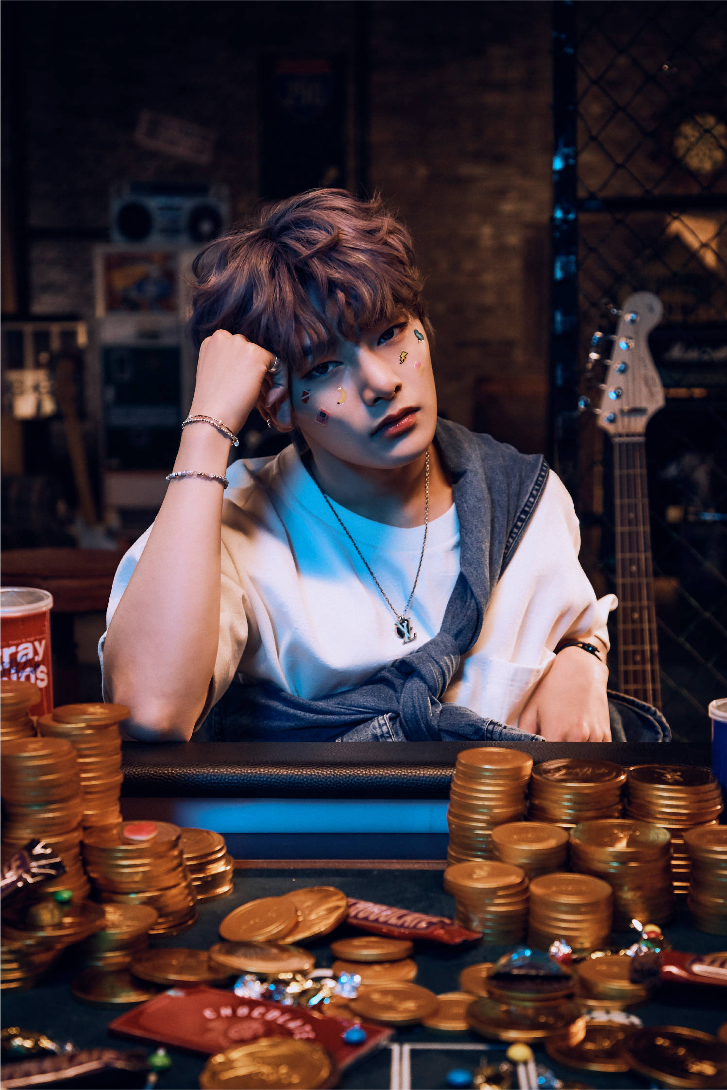 Stray Kids 桌布 壁紙 — 一名年輕男子臉上塗有顏料，疲倦地靠在一張堆滿金幣和糖果的桌子上，房間光線昏暗，由磚塊砌成。