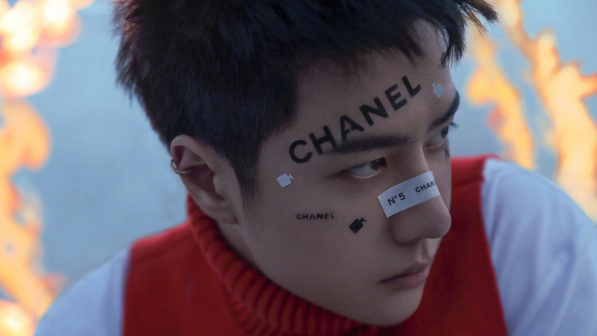 王一博 桌布 壁紙 — 一名年輕男子臉上寫著「CHANEL」和「N°5」，穿著紅色高領毛衣，背景是火熱的橙色和藍色。