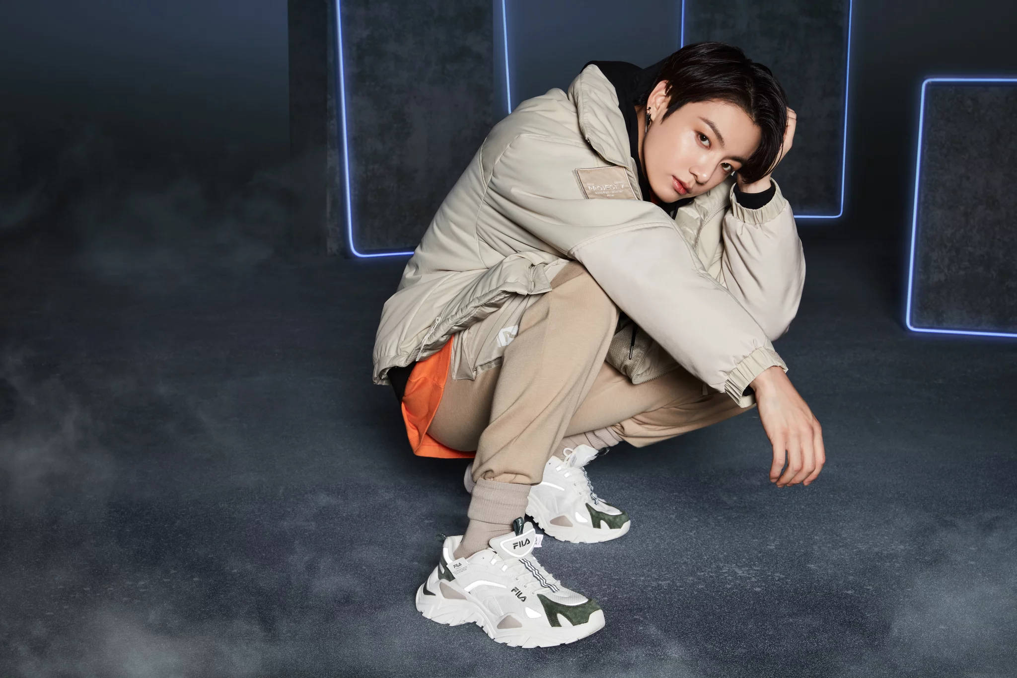 Fila 桌布 壁紙 — 一名年輕男子蹲在煙霧瀰漫的攝影棚中，身穿米色羽絨外套和白色FILA運動鞋，身後是發光的霓虹藍牆壁。