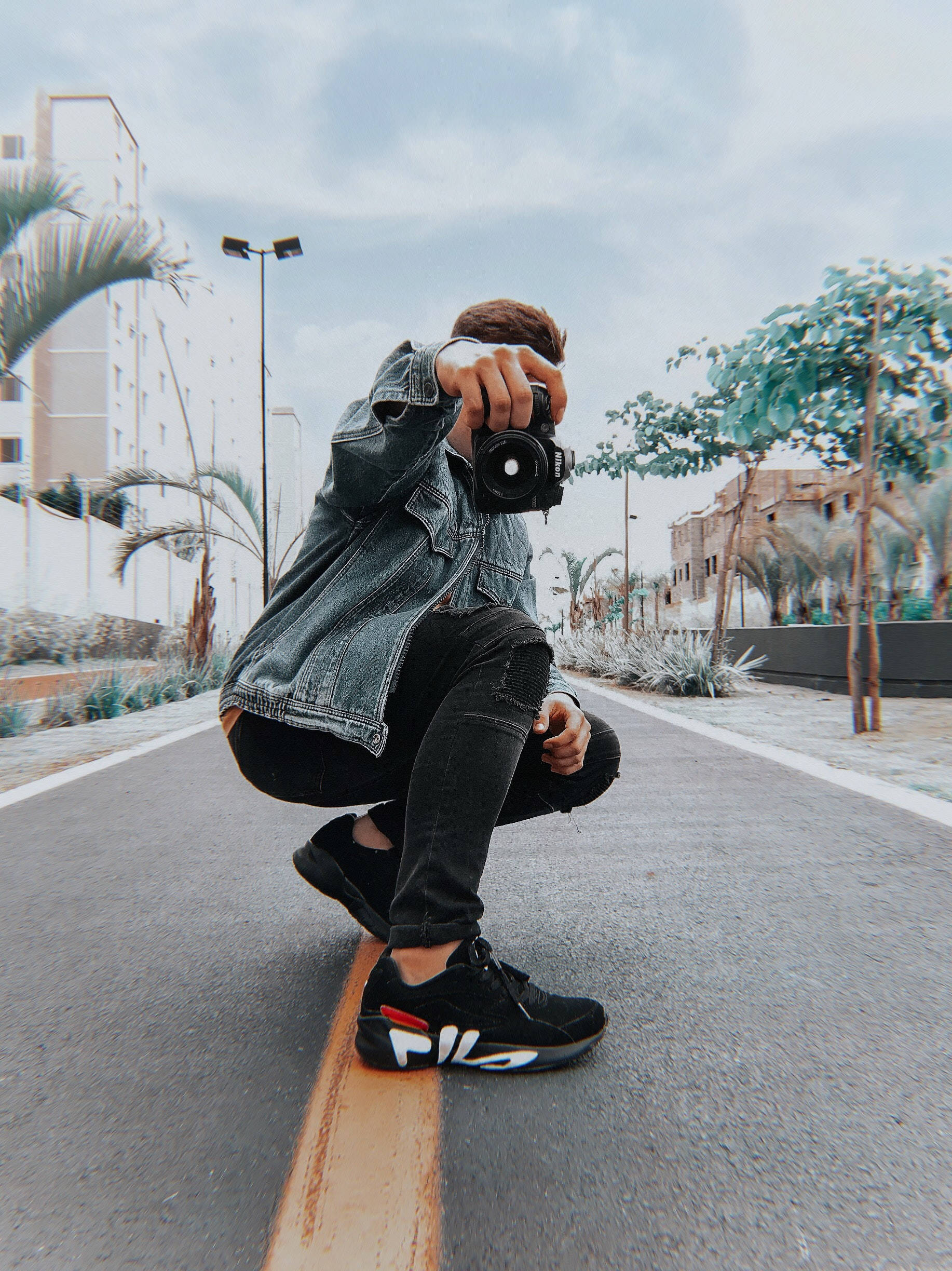 photography iPhone 桌布 壁紙 — 一名年輕男子蹲在鋪砌的道路上，手持相機，身穿牛仔外套和黑色運動鞋。