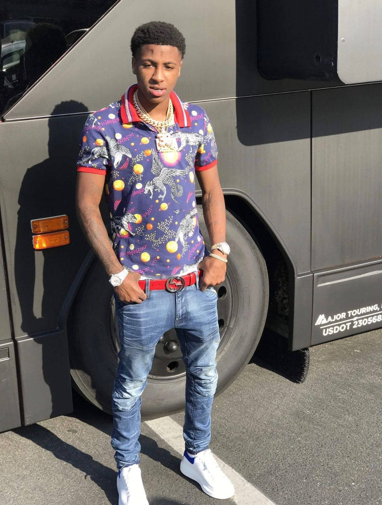 Youngboy 桌布 壁紙 — 一名年輕男子身穿充滿活力的宇宙圖案Polo衫和牛仔褲，在明亮的陽光下自信地站在一輛大型灰色旅遊巴士旁。