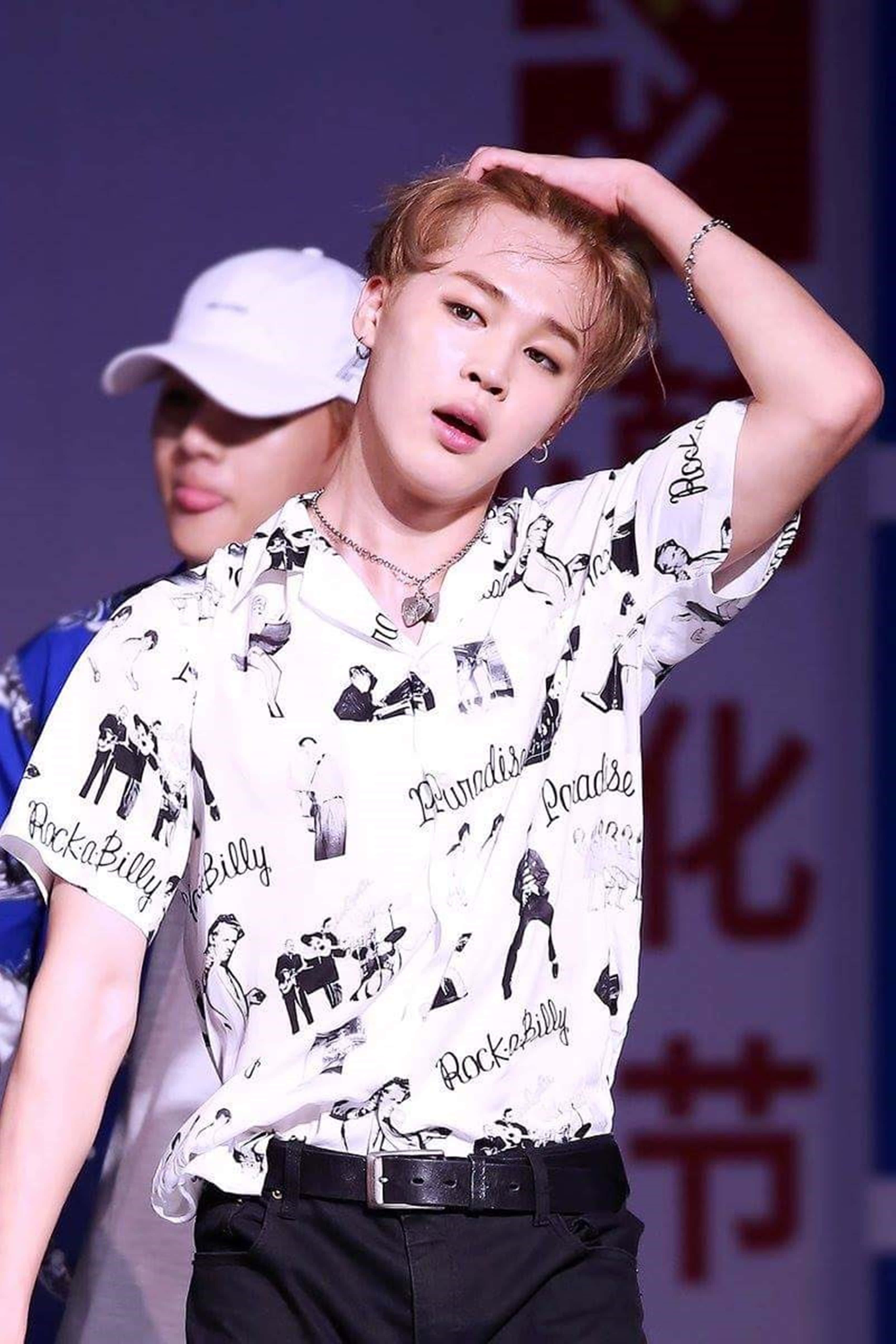 Bts Jimin 桌布 壁紙 — 一名年輕男子身穿白色搖滾印花襯衫和黑色褲子，在舞台燈光下將手放在頭髮上擺姿勢。