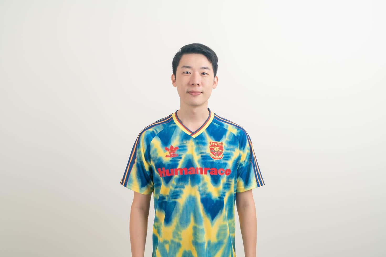 扎染 桌布 壁紙 — 一名年輕男子身穿鮮豔的藍黃紮染足球球衣，上面有紅色文字，站在純白色背景前。