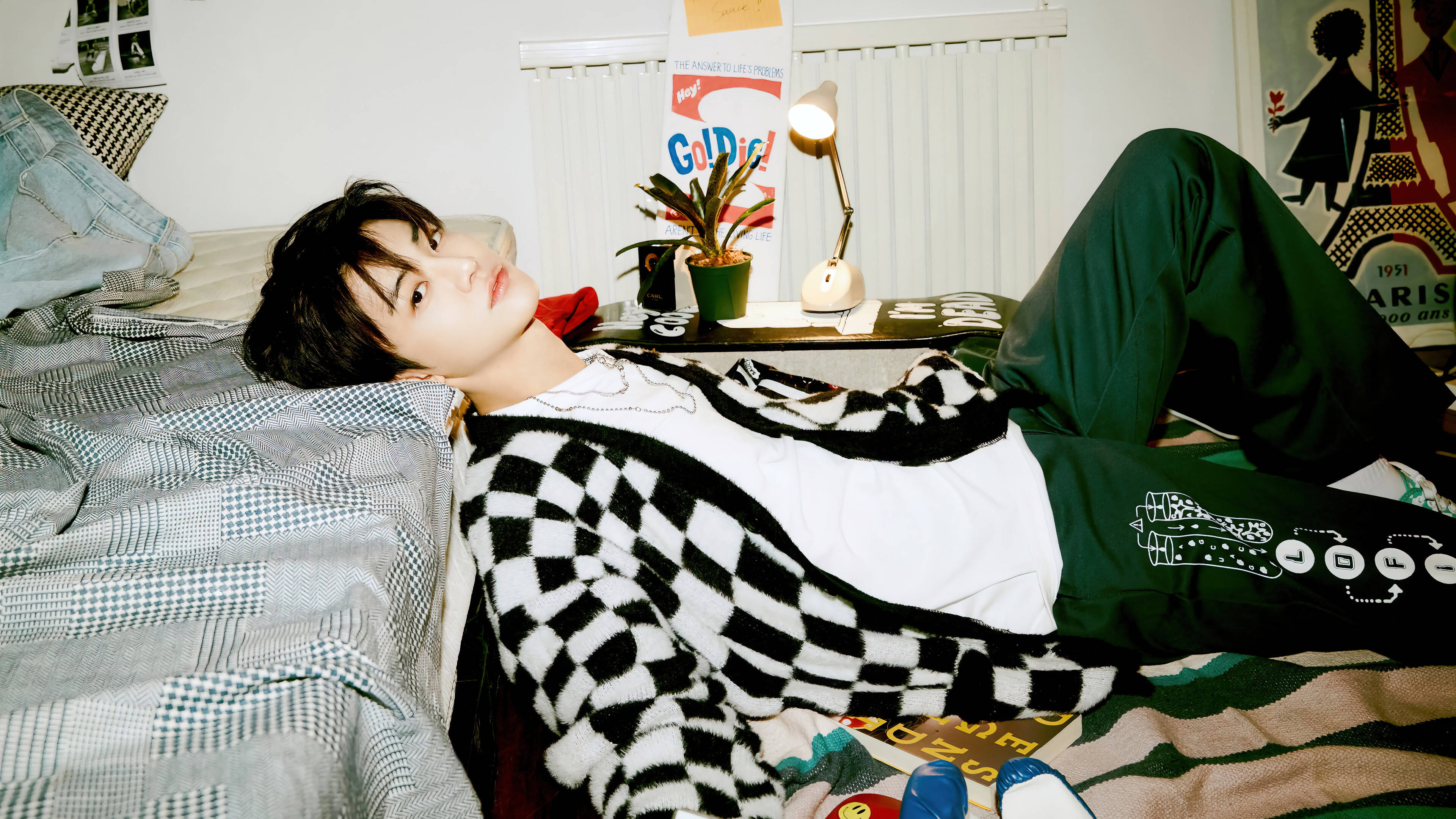 jaemin 桌布 壁紙 — 一名年輕男子身穿黑白格紋開襟毛衣和綠色褲子，隨意地躺在床上，周圍有海報和一盞亮著的檯燈。