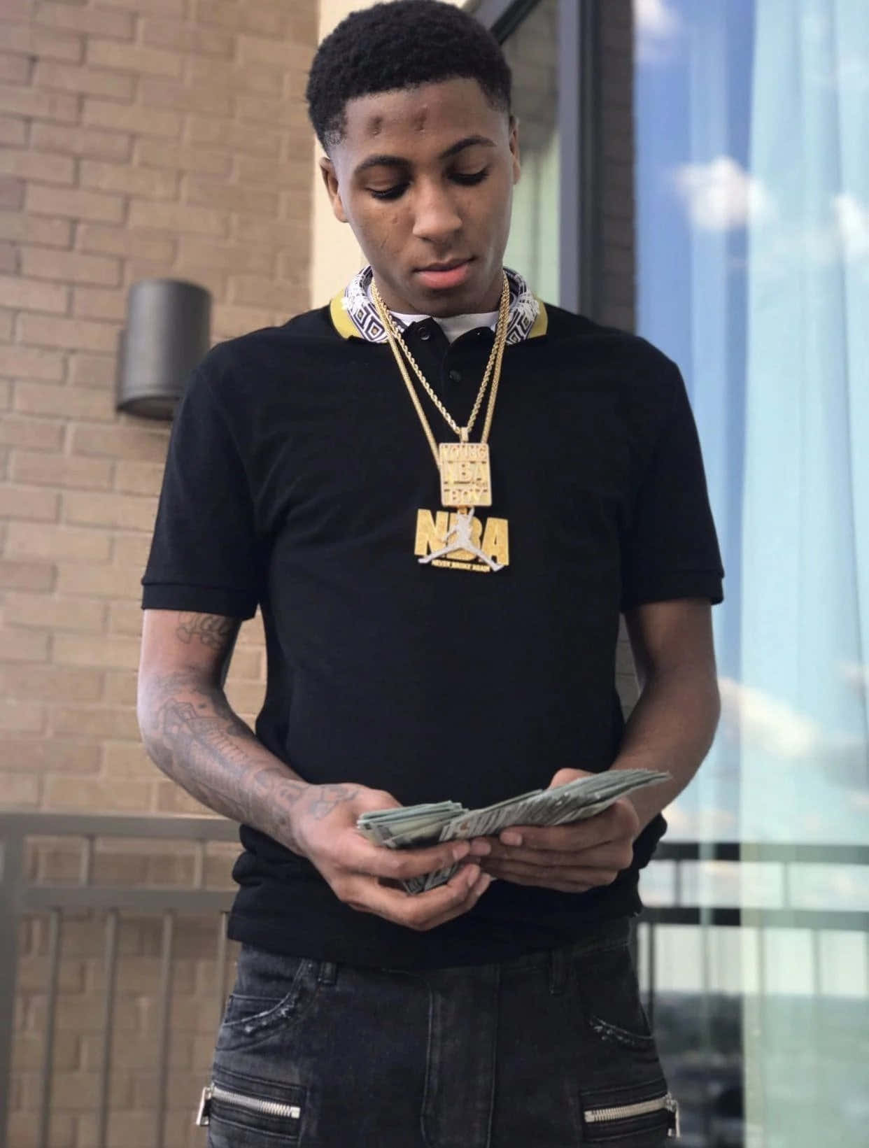 NBA YoungBoy 桌布 壁紙 — 一名年輕男子身穿黑色NBA Polo衫和金色鏈條，站在磚砌建築外數錢。
