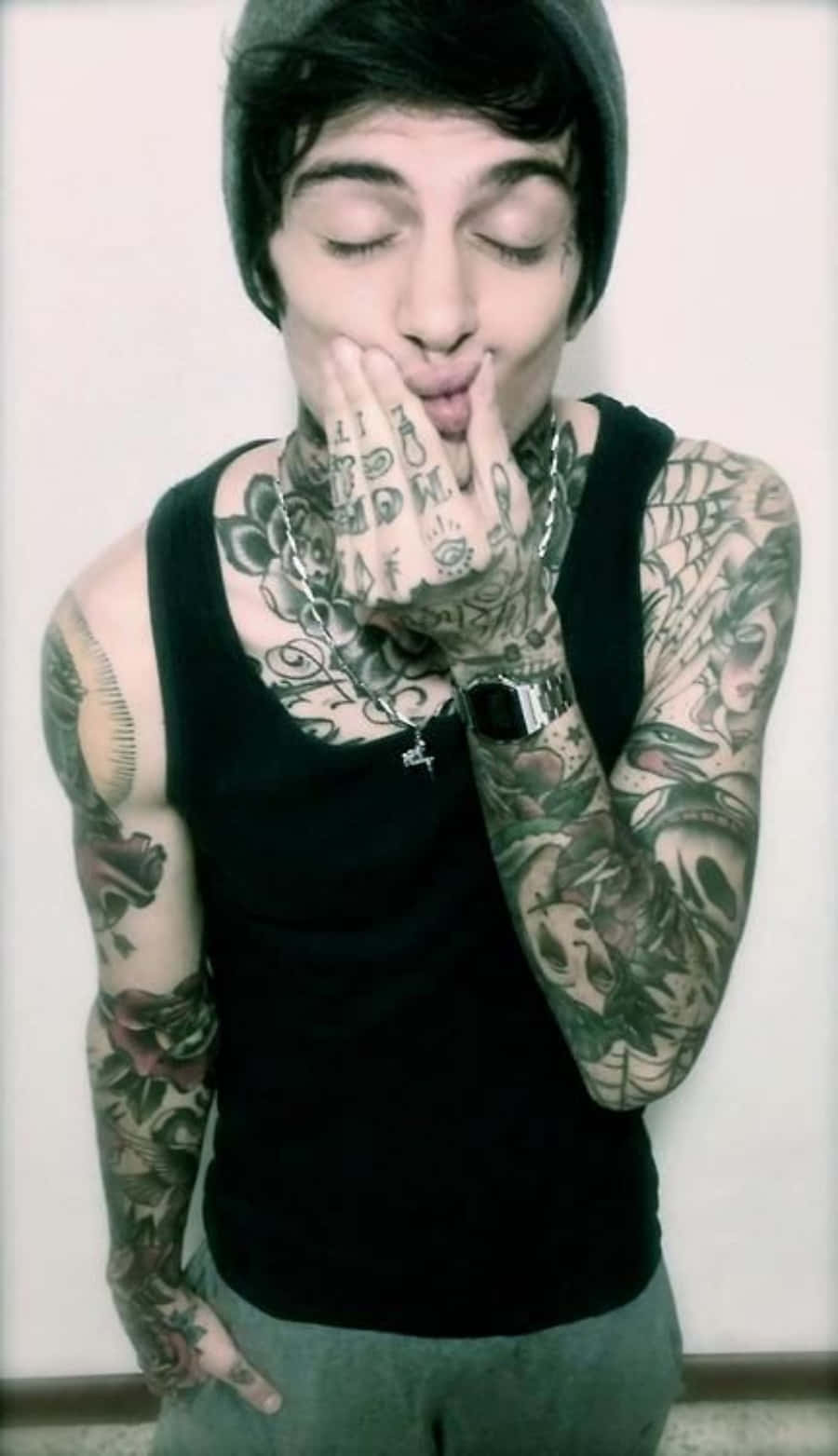 Tattoo Boy 桌布 壁紙 — 一名年輕男子閉上眼睛，雙手靠近嘴巴，在素色牆壁前擺姿勢，展示覆蓋手臂和頸部的大量黑色和灰色刺青。