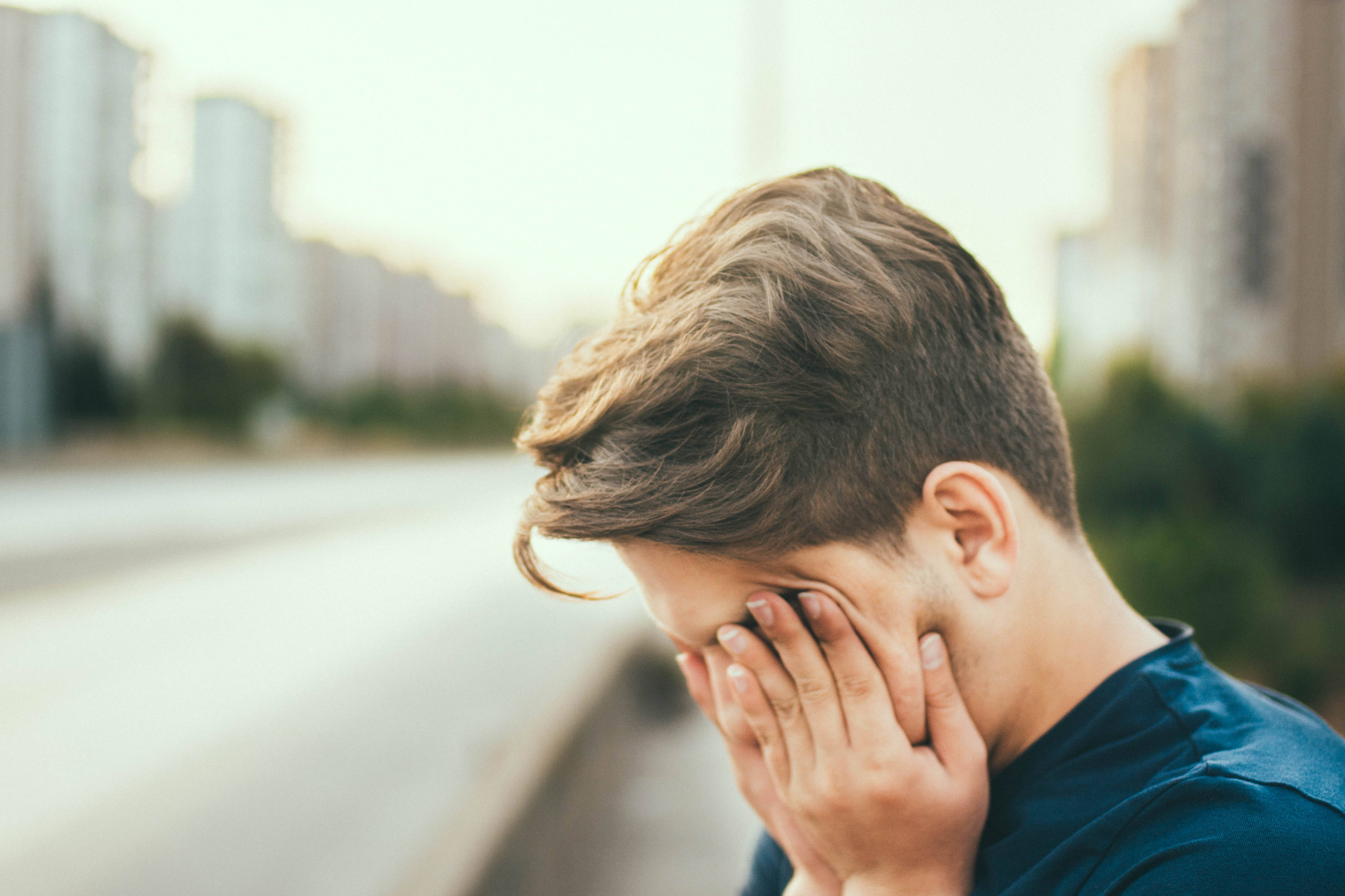 Alone Boy 桌布 壁紙 — 一名年輕男子雙手掩面，獨自站在城市街道上，在模糊的城市背景下傳達深深的痛苦。