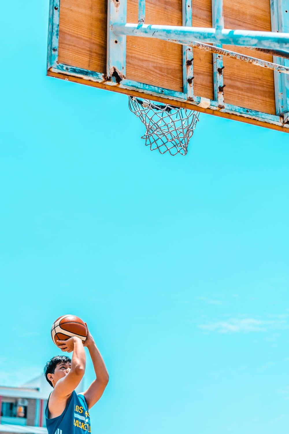 Basketball Aesthetic 桌布 壁紙 — 一名年輕籃球運動員在破舊的籃框下跳起投籃，背景是清澈的藍天。