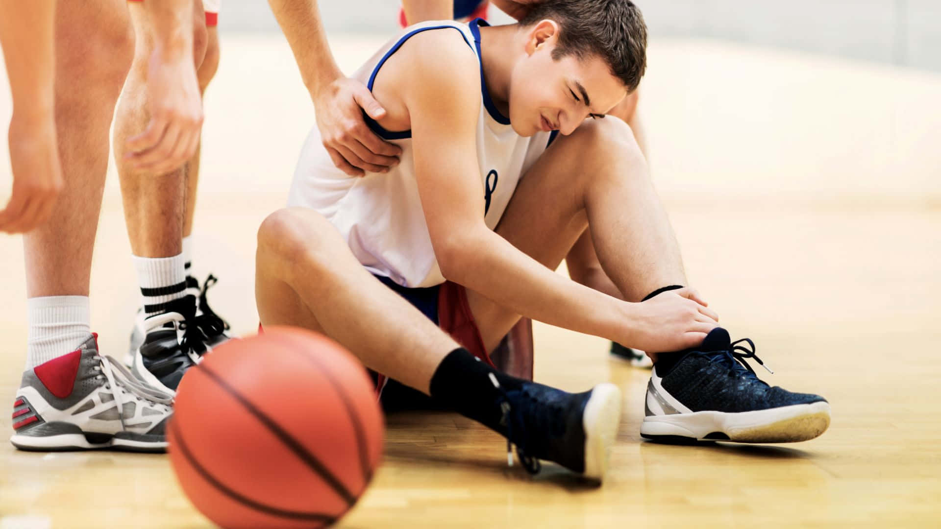 Best Sports 桌布 壁紙 — 一名年輕籃球運動員坐在體育館地板上，痛苦地抓著腳踝，隊友們圍在他身邊。