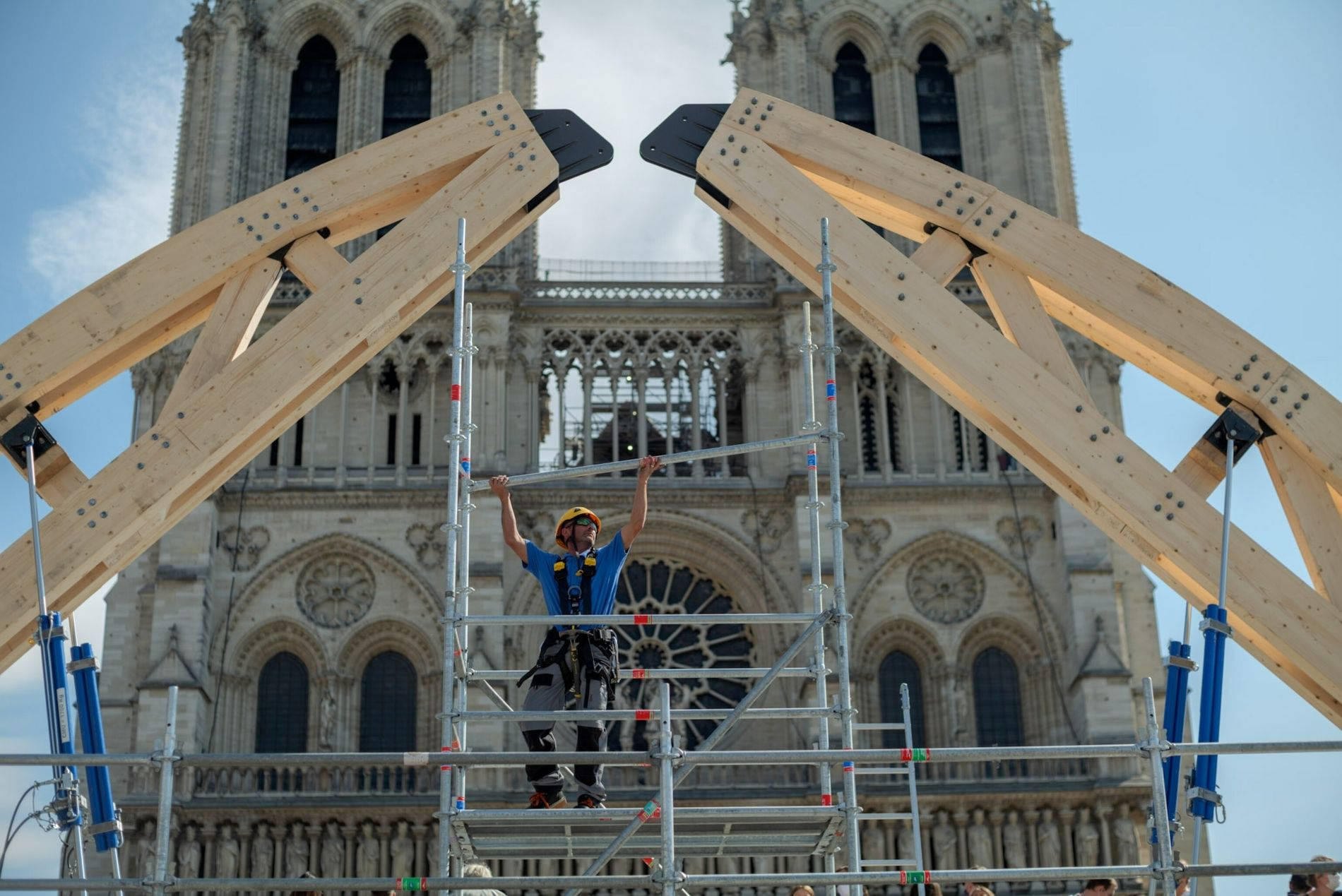 Notre Dame 桌布 壁紙 — 一名建築工人在鷹架上慶祝，巨大的木樑在晴朗的藍天下框住了標誌性的聖母院大教堂。
