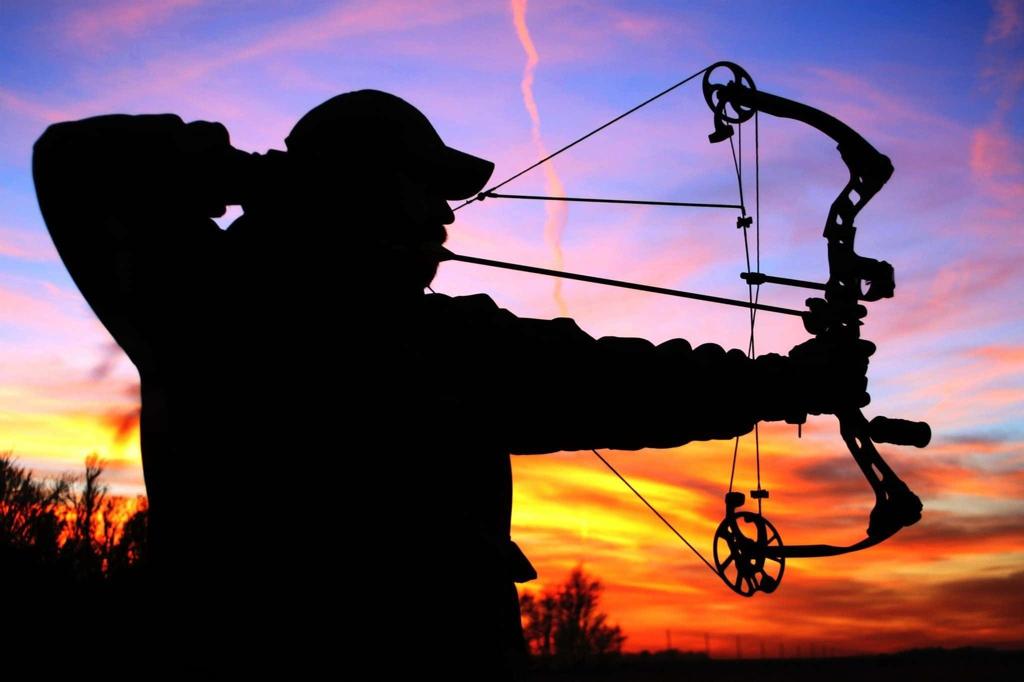 Archery 桌布 壁紙 — 一名弓獵手的剪影，正拉開複合弓，背景是帶有紫色和橙色條紋的熾熱日落天空。
