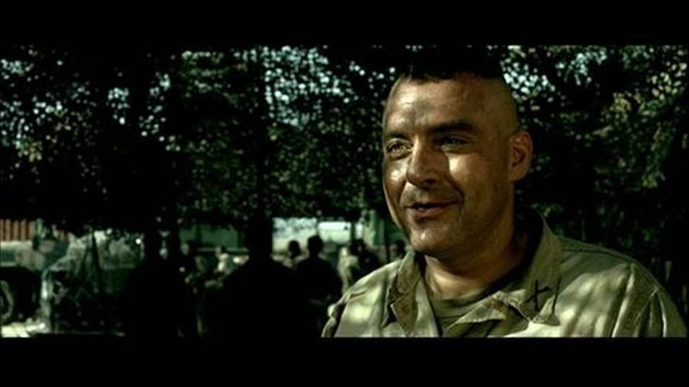 電影 桌布 壁紙 — 一名微笑的光頭士兵穿著軍服，在戶外樹下站立，背景有車輛和人物的特寫。