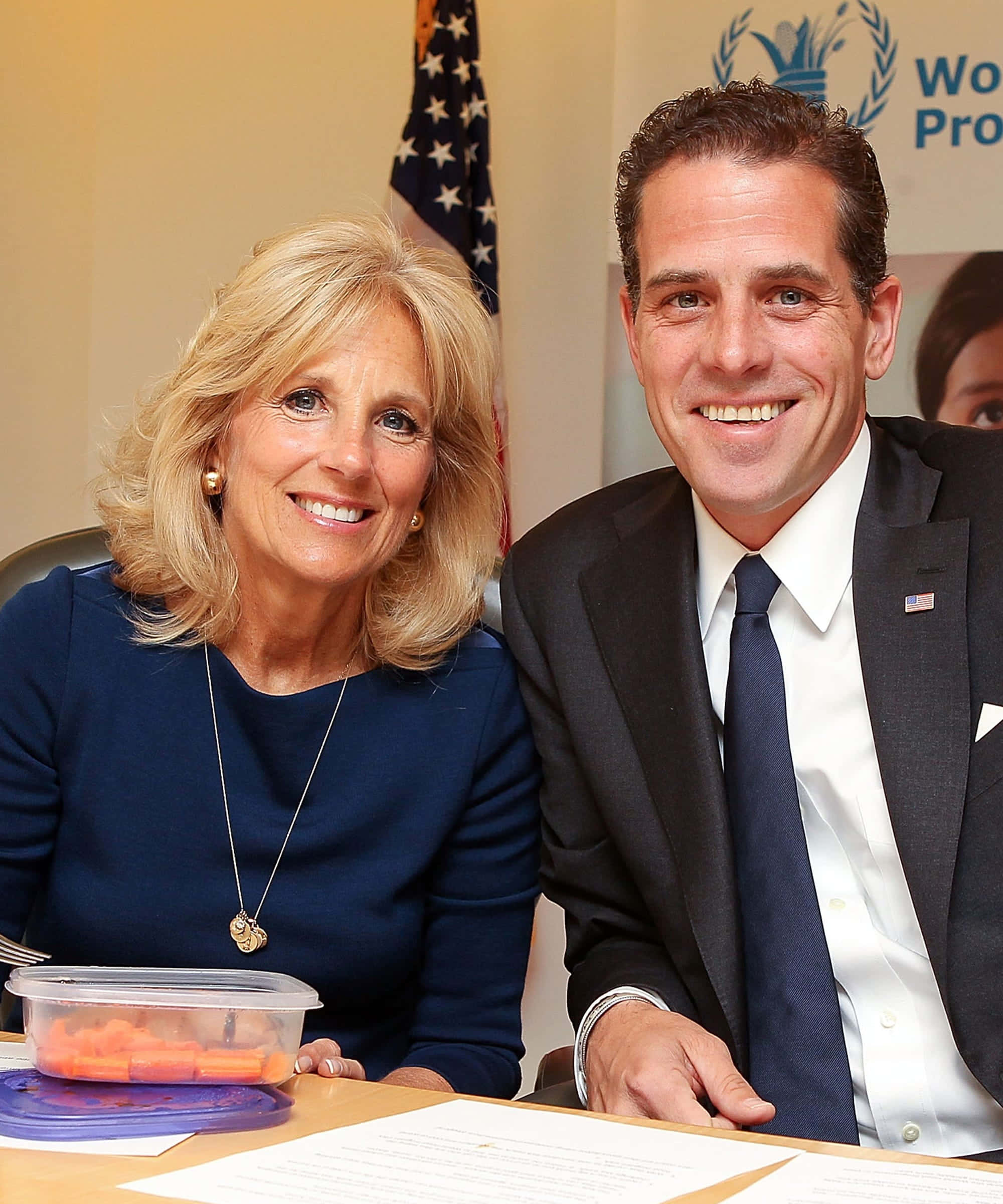 Joe Biden 桌布 壁紙 — 一名微笑的男士和女士穿著正式服裝坐在辦公桌前，桌上放有文件和一個裝有胡蘿蔔條的容器，身後有美國國旗和世界糧食計劃署的橫幅。