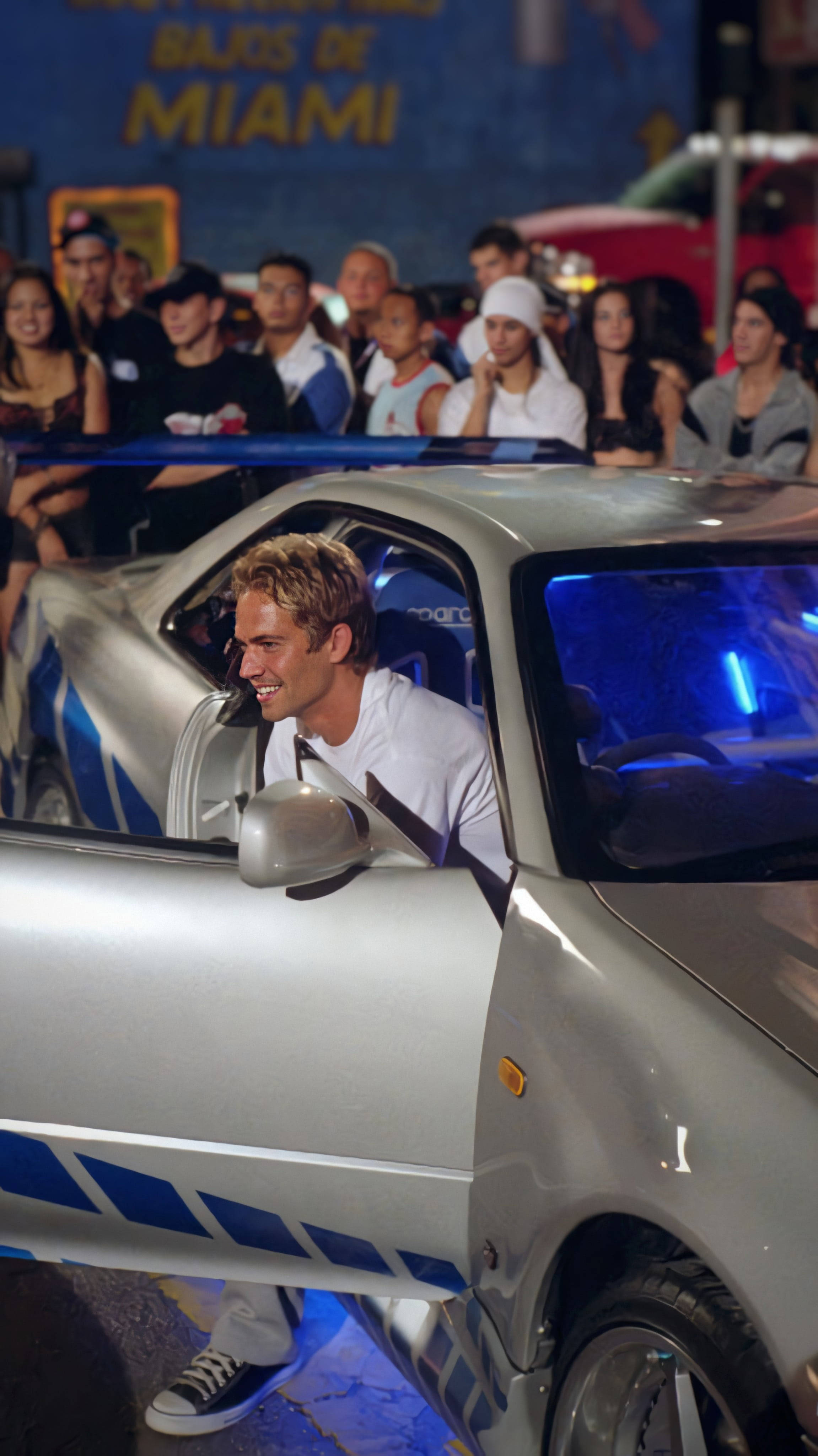 Paul Walker 桌布 壁紙 — 一名微笑的男子從帶有藍色裝飾的銀色賽車中探出身子，夜晚人群在旁觀看。