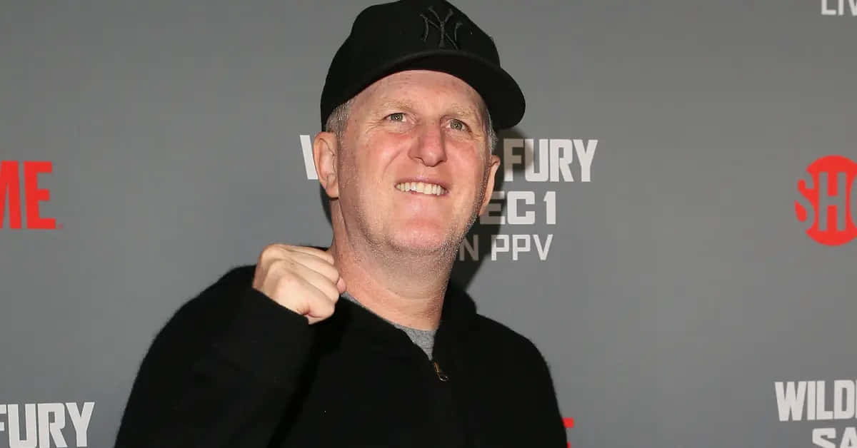 Michael Rapaport 桌布 壁紙 — 一名微笑的男子戴著黑色帽子和穿著黑色外套，在灰色活動背景前握拳擺姿勢，背景有紅色和白色的標誌。