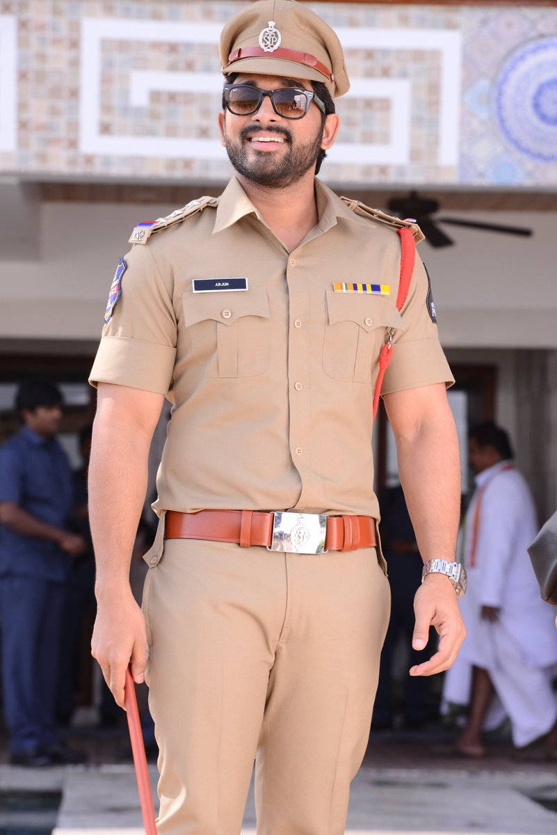 Allu Arjun 桌布 壁紙 — 一名微笑的男子穿著卡其色警察制服，戴著太陽眼鏡，手持紅色警棍，自信地站在戶外。