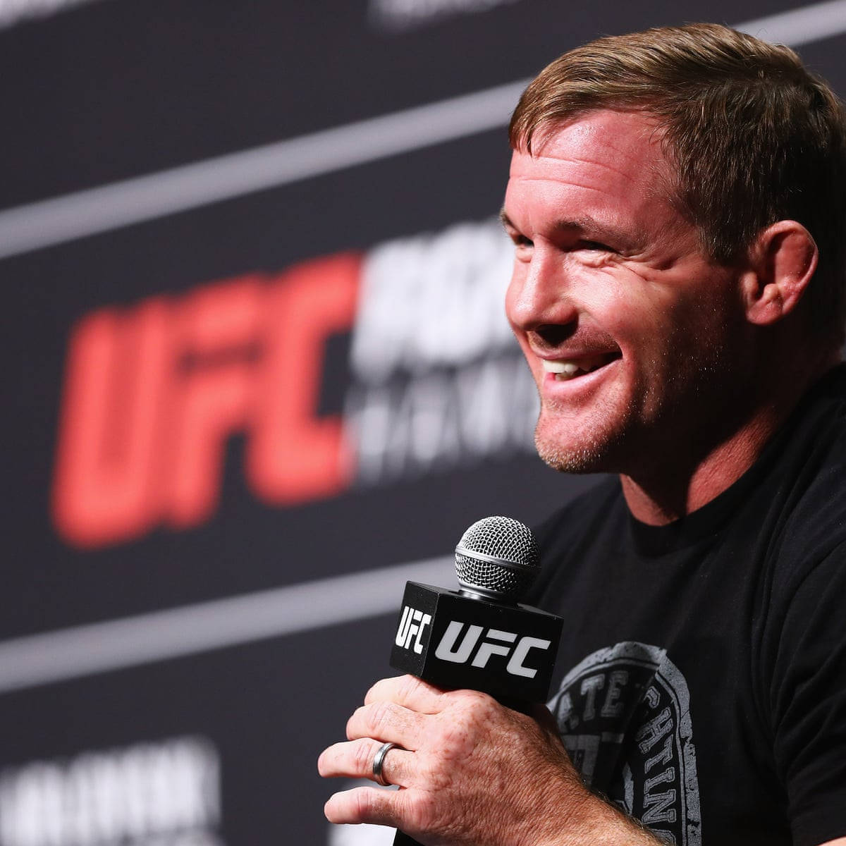 UFC 桌布 壁紙 — 一名微笑的男子穿著黑色UFC T恤，在品牌背景的記者會上拿著UFC麥克風。