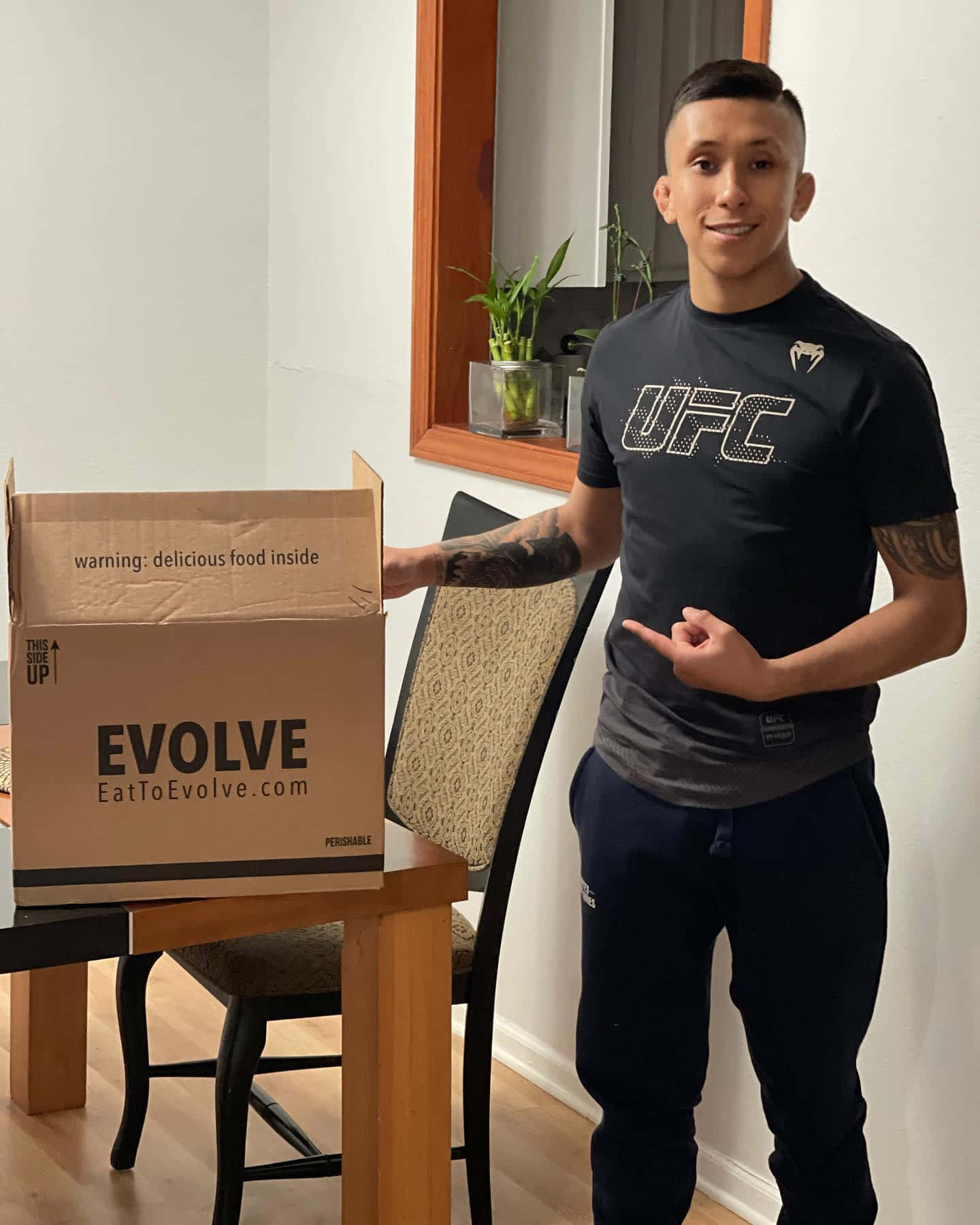 Fitness 桌布 壁紙 — 一名微笑的男子身穿UFC襯衫，站在一個標有「EVOLVE」的紙箱旁，上面有開玩笑的警告，說裡面有美味的食物。