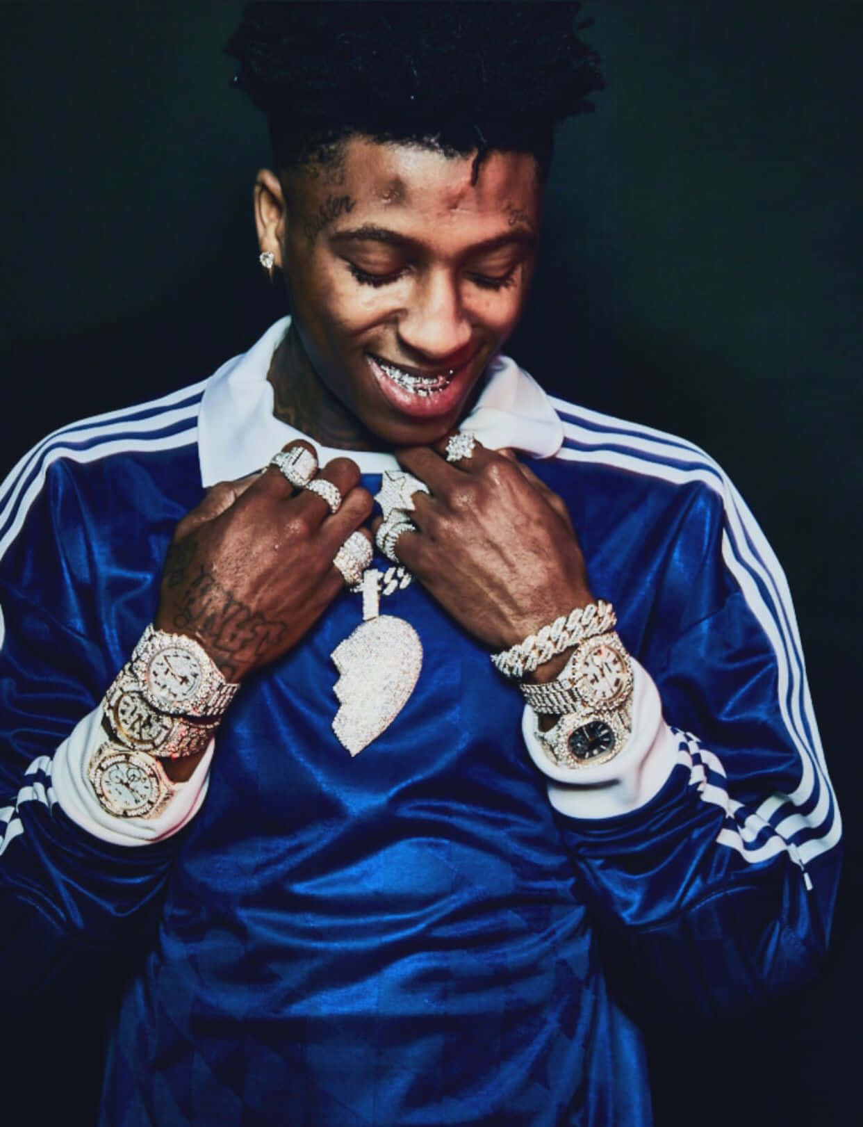 NBA YoungBoy 桌布 壁紙 — 一名微笑的男子身穿藍色天鵝絨運動服，在深色背景前展示多枚鑲鑽手錶和戒指。