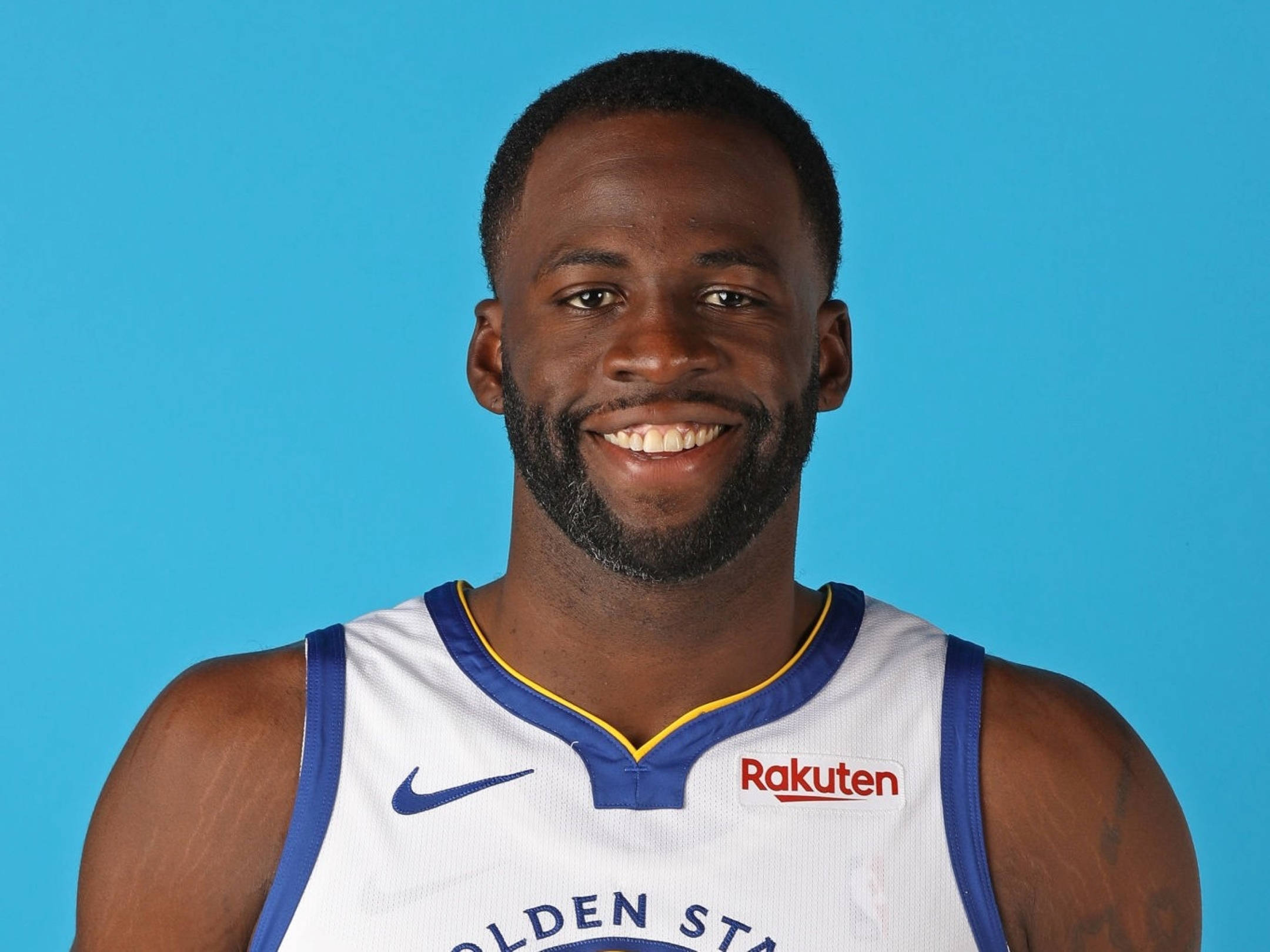 Draymond Green 桌布 壁紙 — 一名微笑的籃球運動員，身穿白色金州勇士隊球衣，背景為純藍色。
