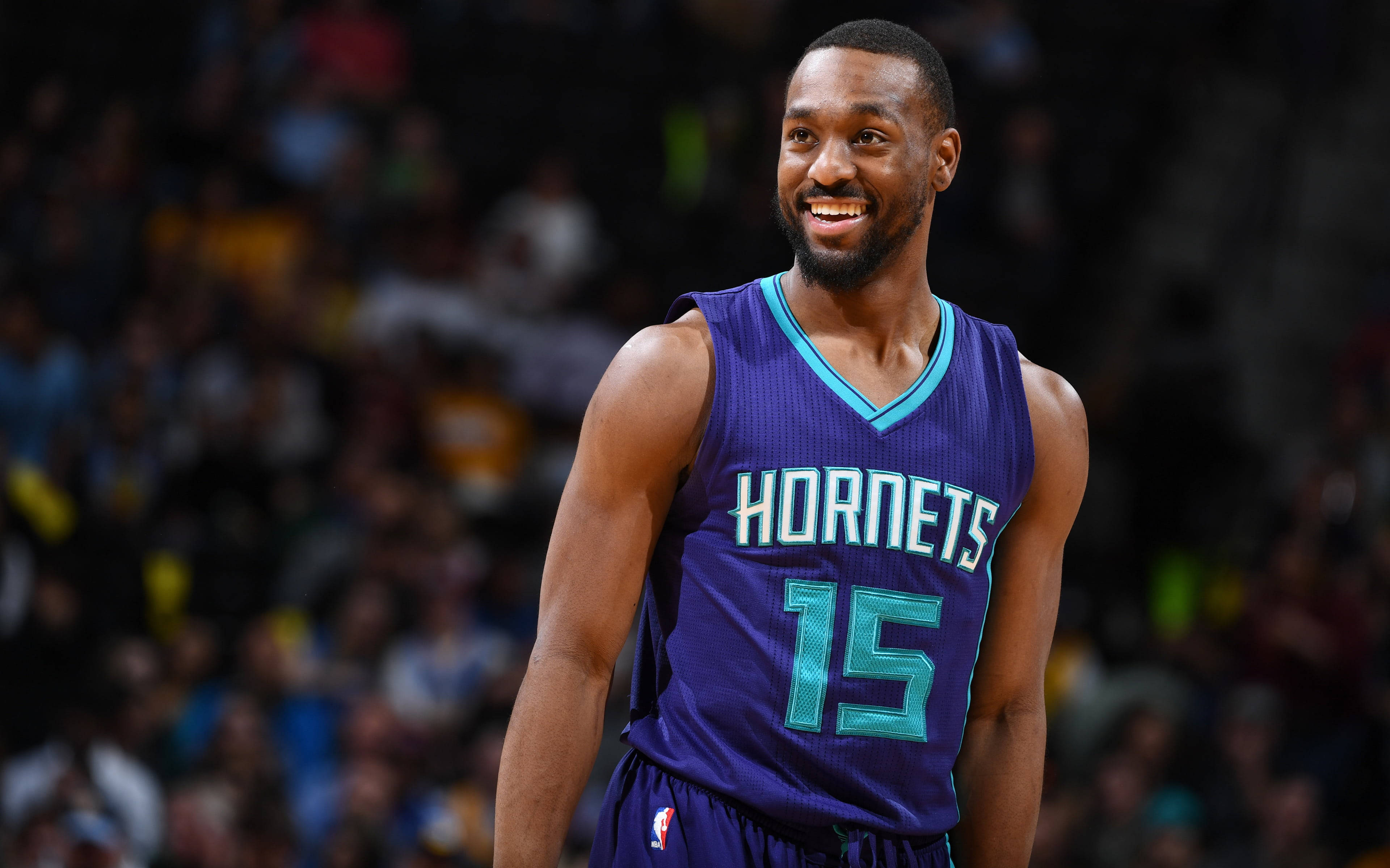 Kemba Walker 桌布 壁紙 — 一名微笑的籃球運動員身穿紫色黃蜂隊15號球衣，自信地站在球場上，背景是模糊的觀眾。