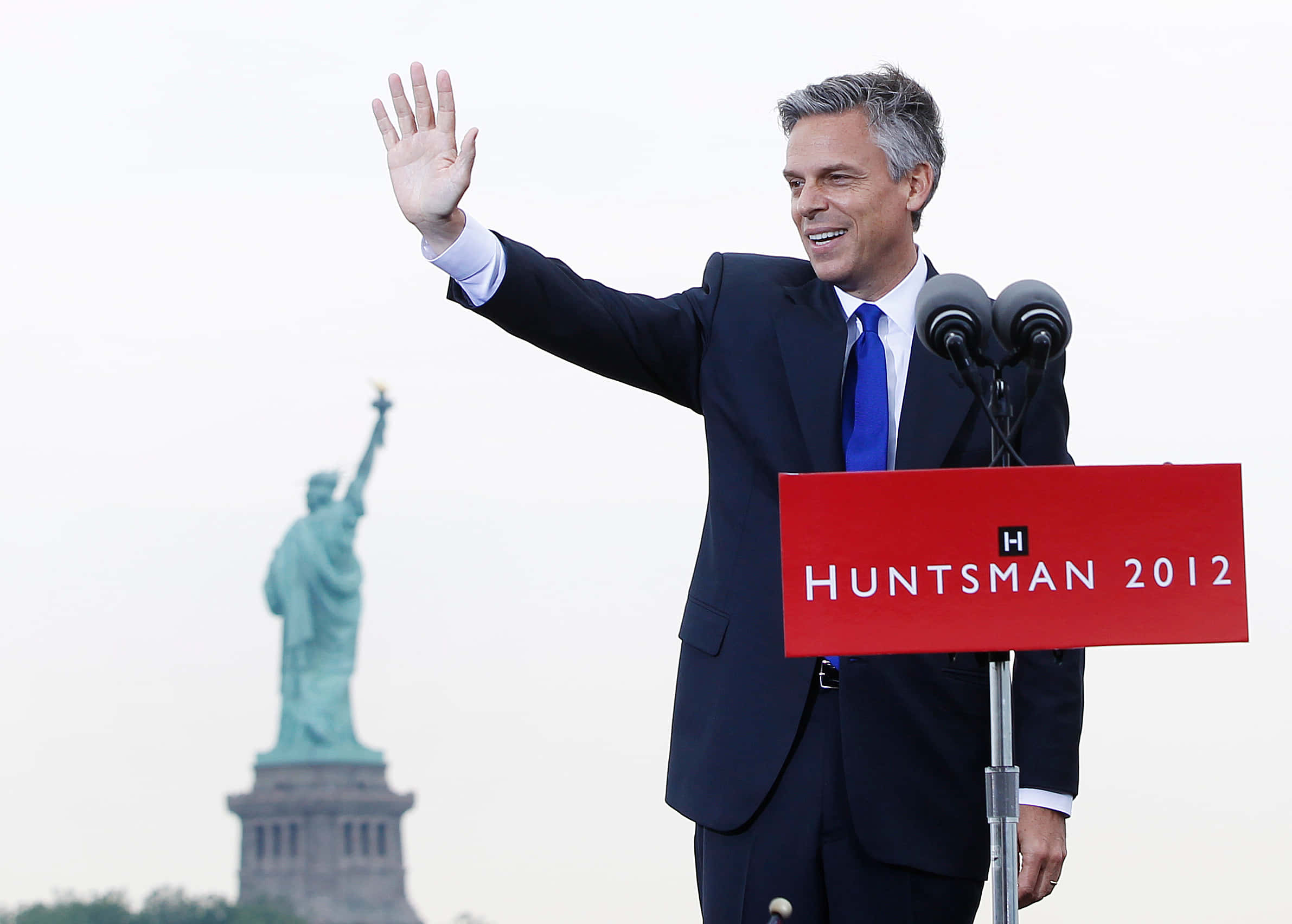 桌布 壁紙 — 一名微笑的西裝男子在標有 Huntsman 2012 的講台上揮手，背景是自由女神像。