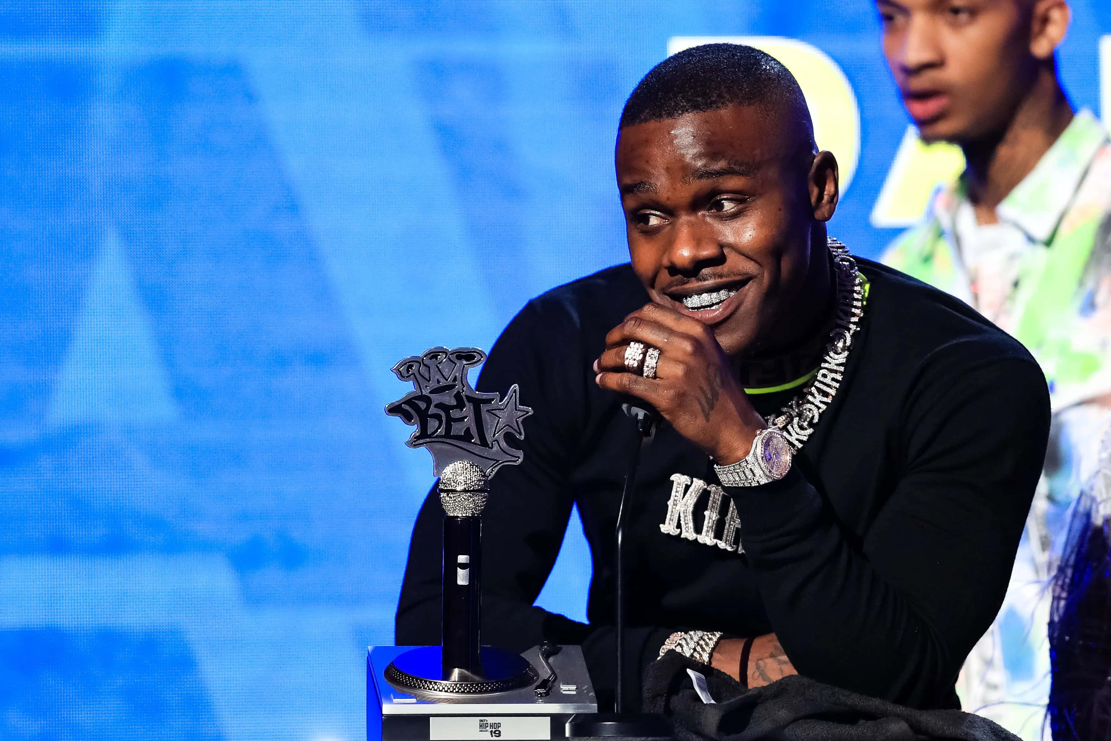 Dababy 桌布 壁紙 — 一名微笑的黑襯衫男子坐在講台上，面前有BET獎項麥克風和獎盃，背景為藍色數位畫面。