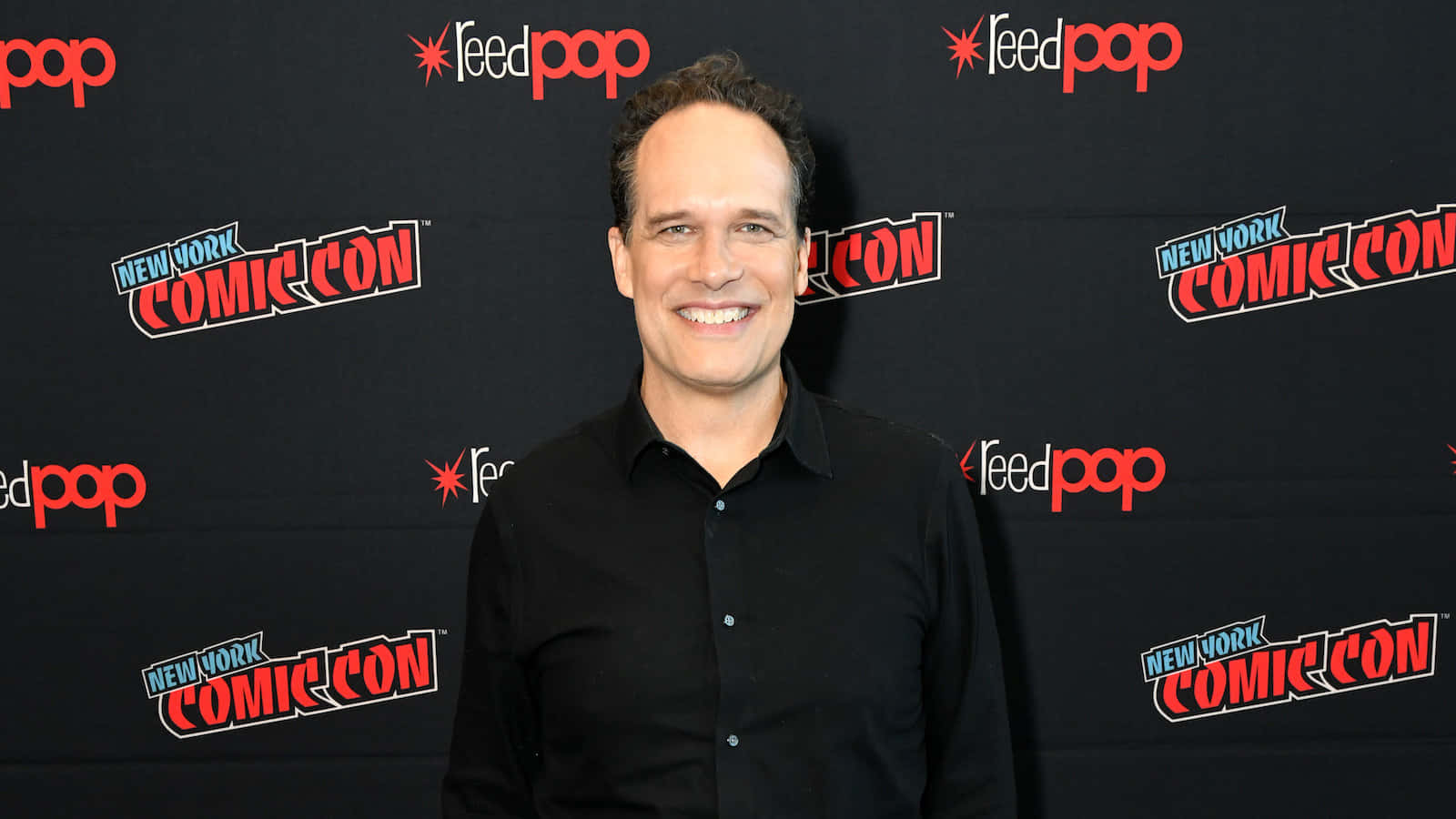 Diedrich Bader 桌布 壁紙 — 一名微笑的黑襯衫男子站在黑色背景前，背景上重複出現紐約動漫展和Feedpop標誌。