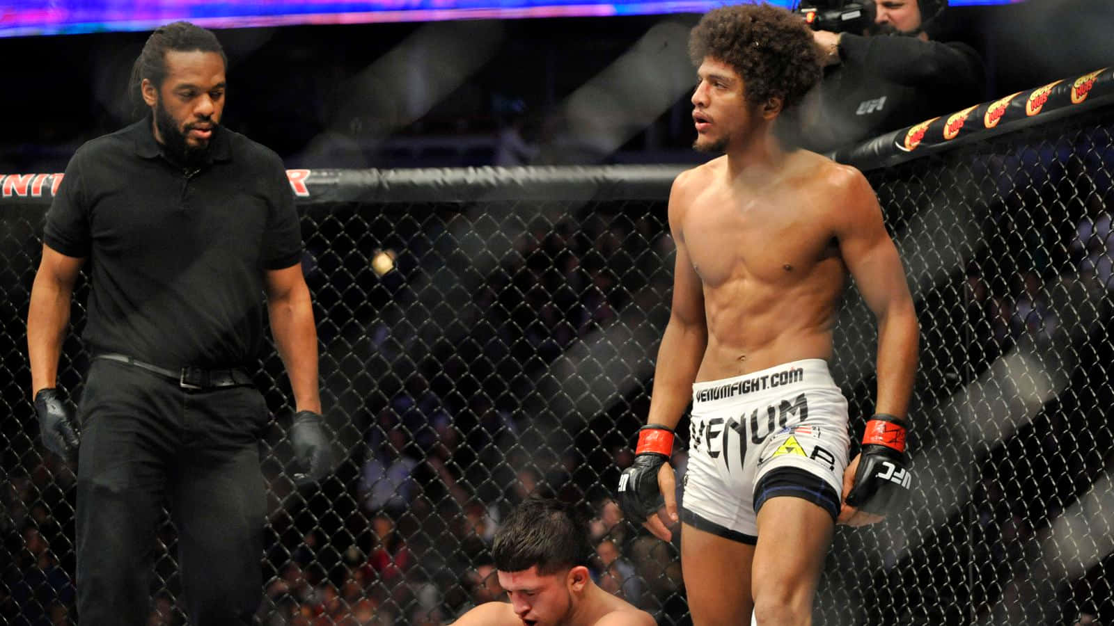 UFC 桌布 壁紙 — 一名戰士在八角籠中勝利站立，對手倒地敗北，裁判在旁密切注視。