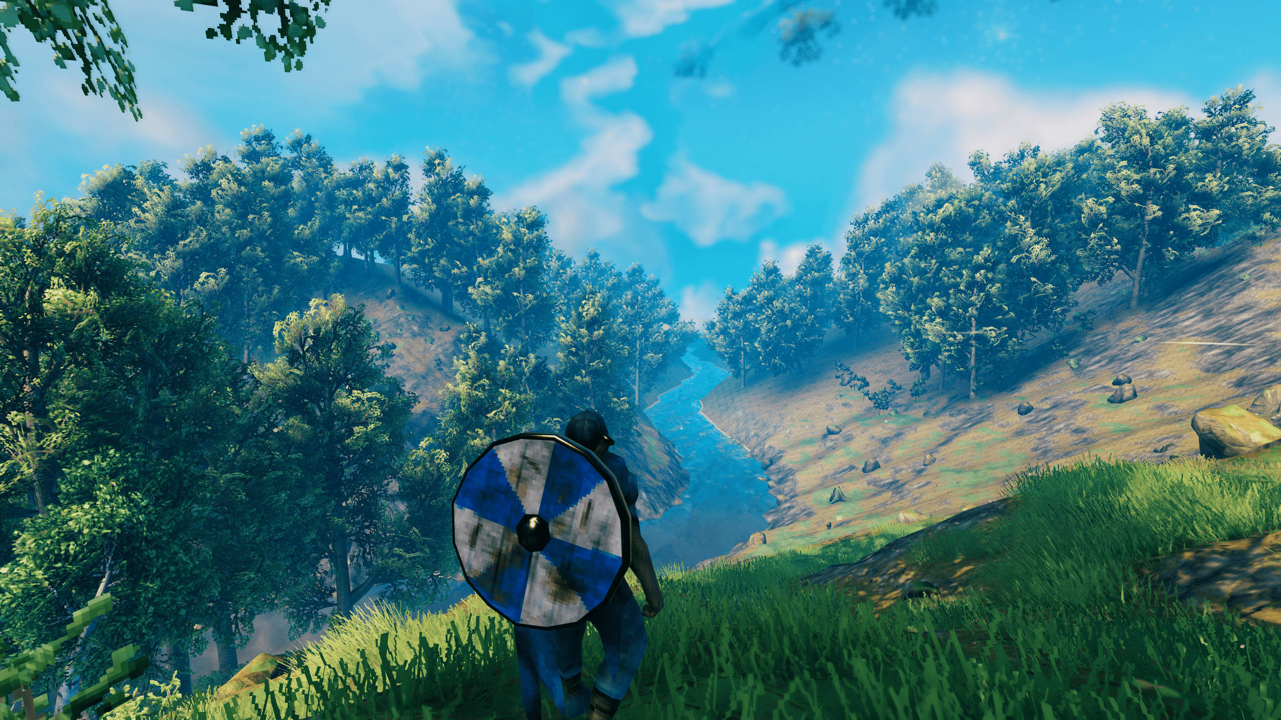 Valheim 桌布 壁紙 — 一名戰士手持藍白盾牌，走過陽光明媚的草地山坡，俯瞰蜿蜒的河流，兩旁是茂密的森林。
