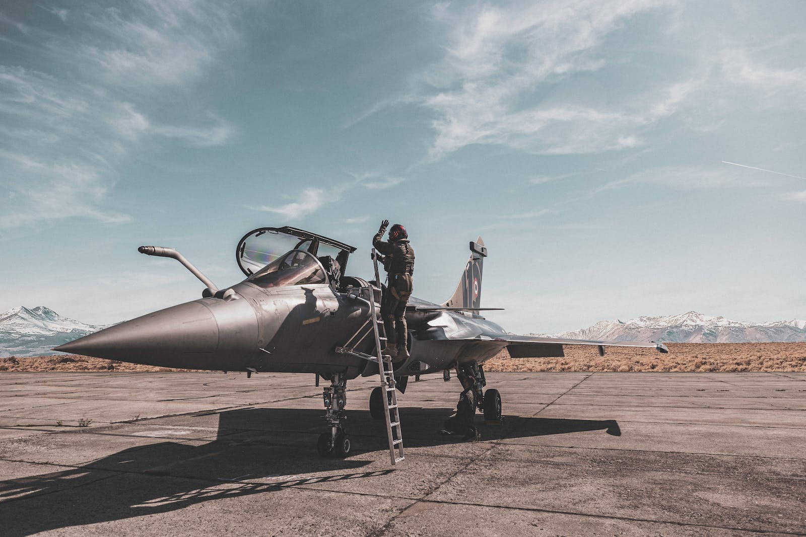 Air Force 桌布 壁紙 — 一名戰鬥機飛行員爬上梯子，進入一架停在停機坪上的流線型噴射機，背景是積雪的山脈，天空部分多雲。
