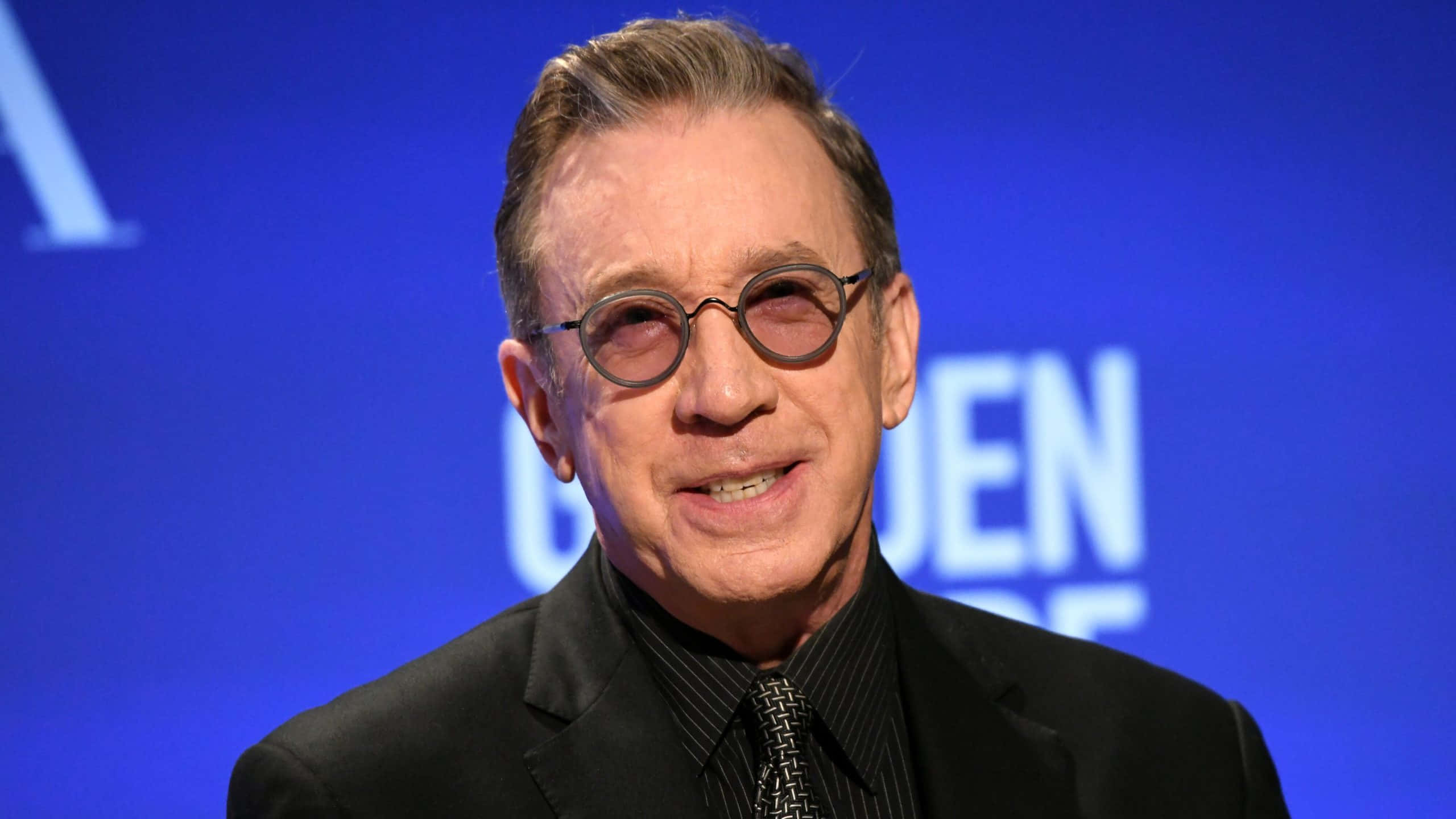 Tim Allen 桌布 壁紙 — 一名戴圓框眼鏡、身穿黑色西裝的男士特寫肖像，背景為藍色，帶有部分白色字樣。