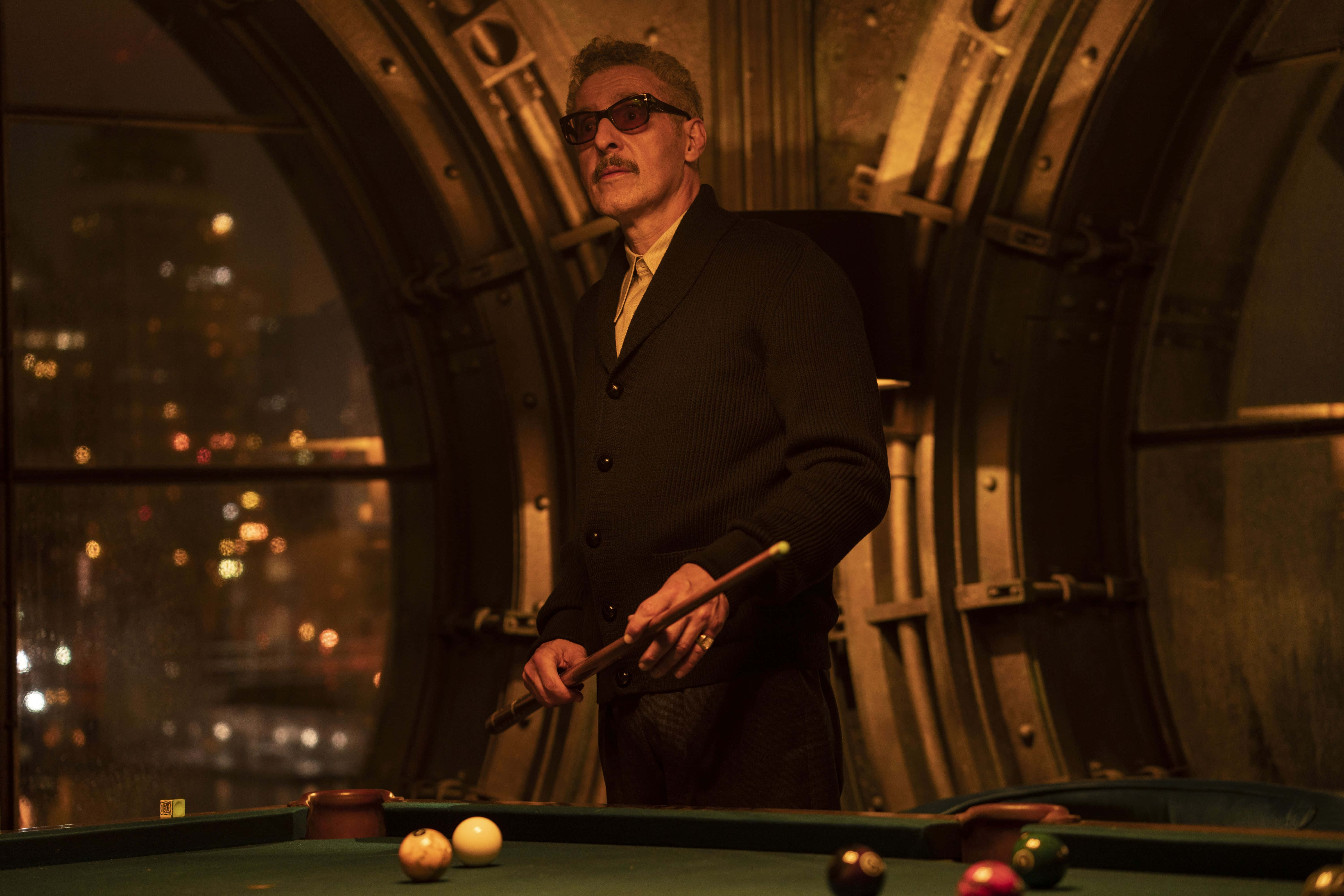 John Turturro 桌布 壁紙 — 一名戴太陽眼鏡、身穿黑色西裝的男子，在光線昏暗的房間裡，手持撞球桿，站在綠色球檯旁，透過大型拱形窗戶可看到城市景觀。