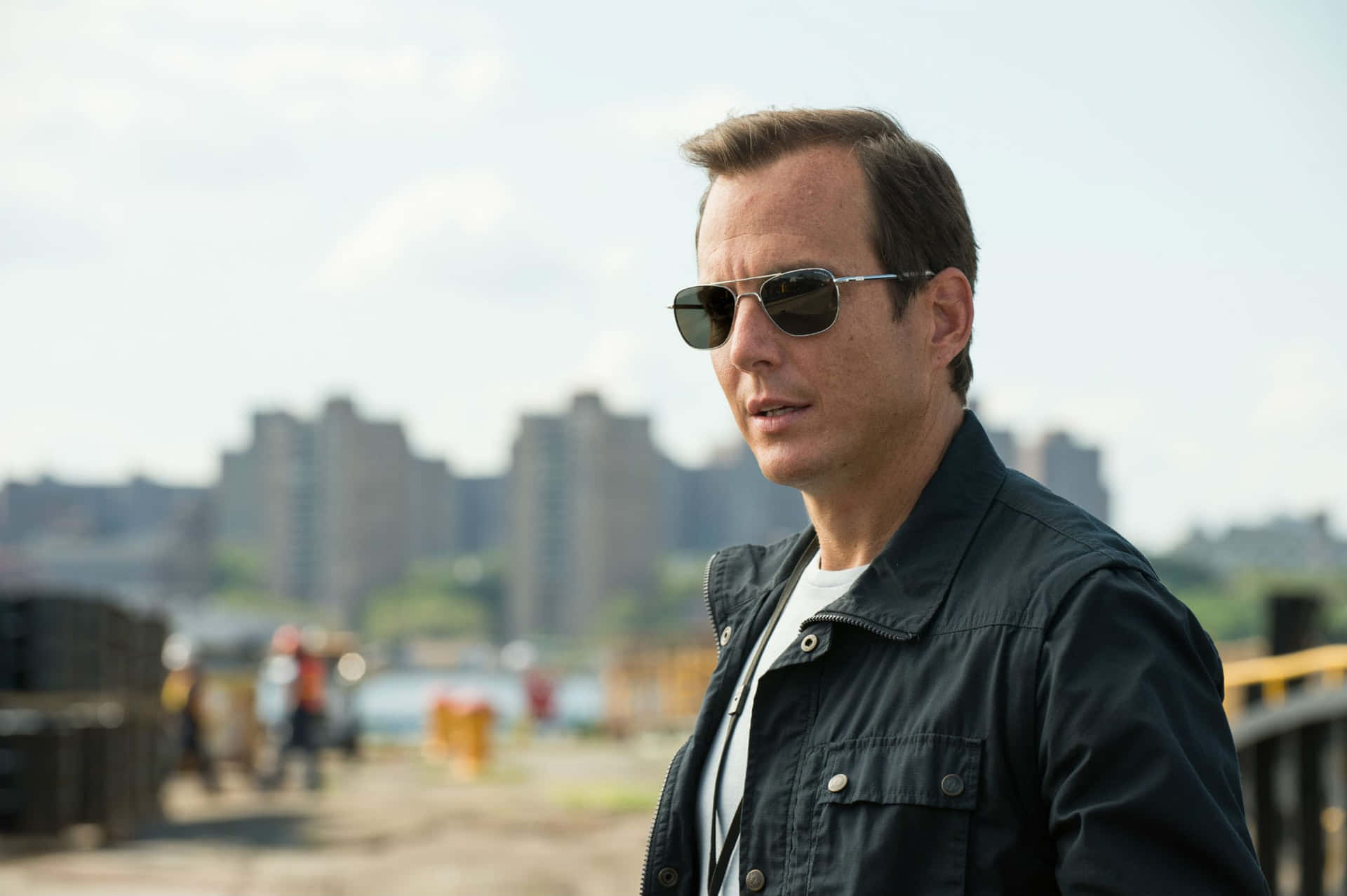 Will Arnett 桌布 壁紙 — 一名戴太陽眼鏡和深色外套的男子站在橋上，俯瞰著晴朗天空下的城市景觀。