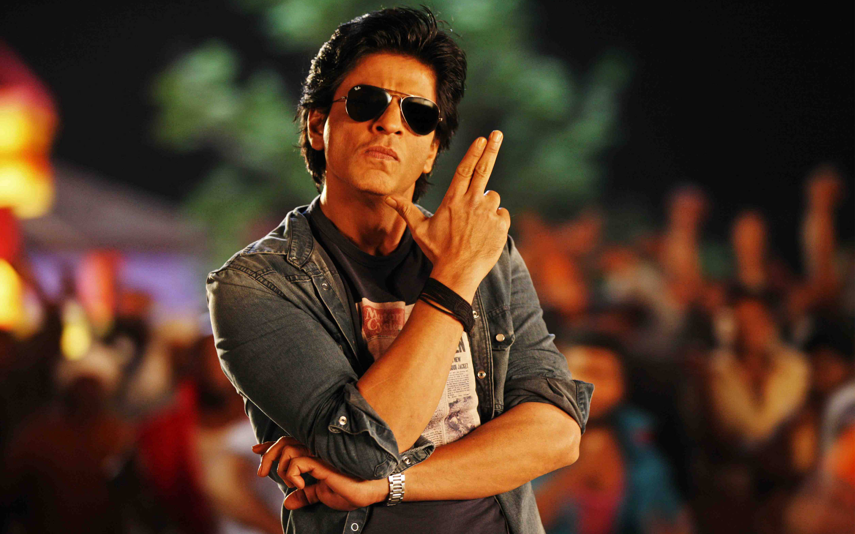 Shahrukh 桌布 壁紙 — 一名戴太陽眼鏡和牛仔外套的男子在夜晚做出和平手勢，身後有一群人。