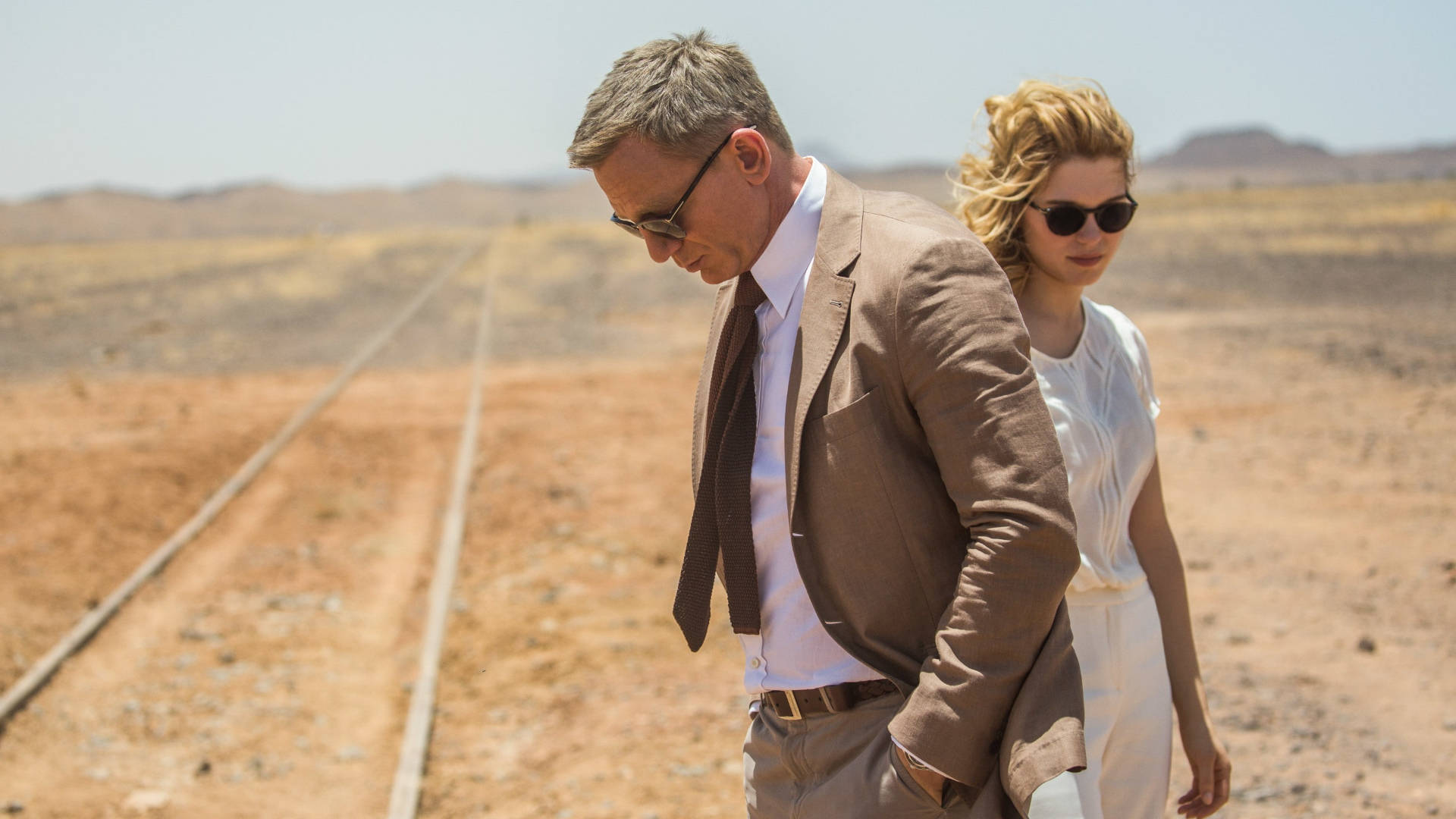 Daniel Craig 桌布 壁紙 — 一名戴太陽眼鏡的男人和女人在烈日下沿著沙漠鐵路軌道行走。