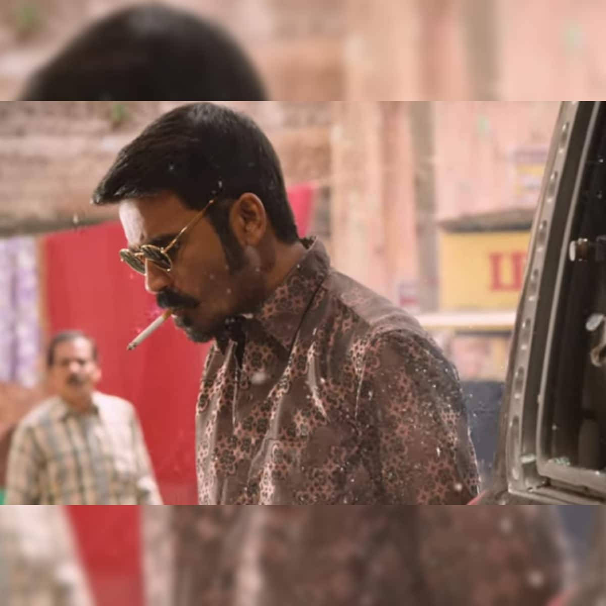 Dhanush 桌布 壁紙 — 一名戴太陽眼鏡的男子在塵土飛揚的城市街道上，在車旁抽煙，背景人物模糊。