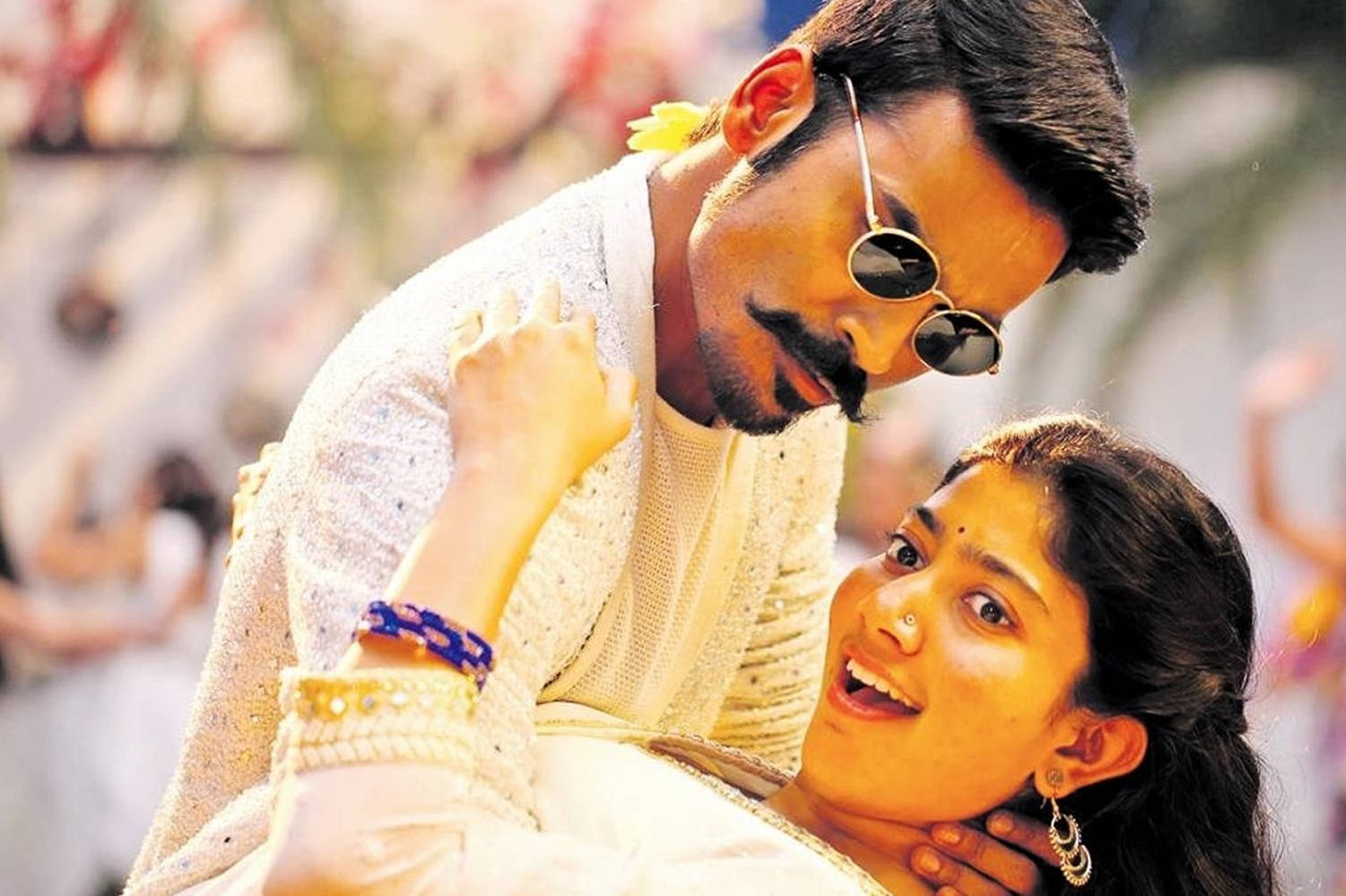 Dhanush 桌布 壁紙 — 一名戴太陽眼鏡的男子在戶外節慶慶祝活動中，俯身靠近一位身穿白色傳統服飾、面帶微笑的女子。