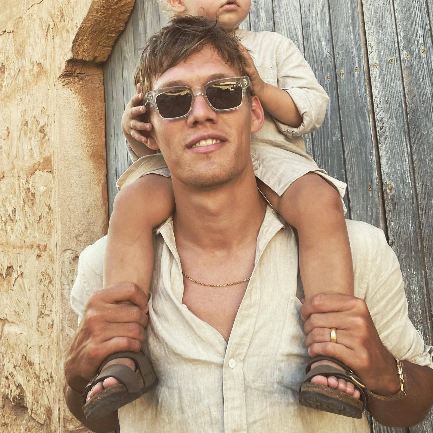 Sunglasses 桌布 壁紙 — 一名戴太陽眼鏡的男子在質樸的石頭和木門背景下，將孩子扛在肩上。