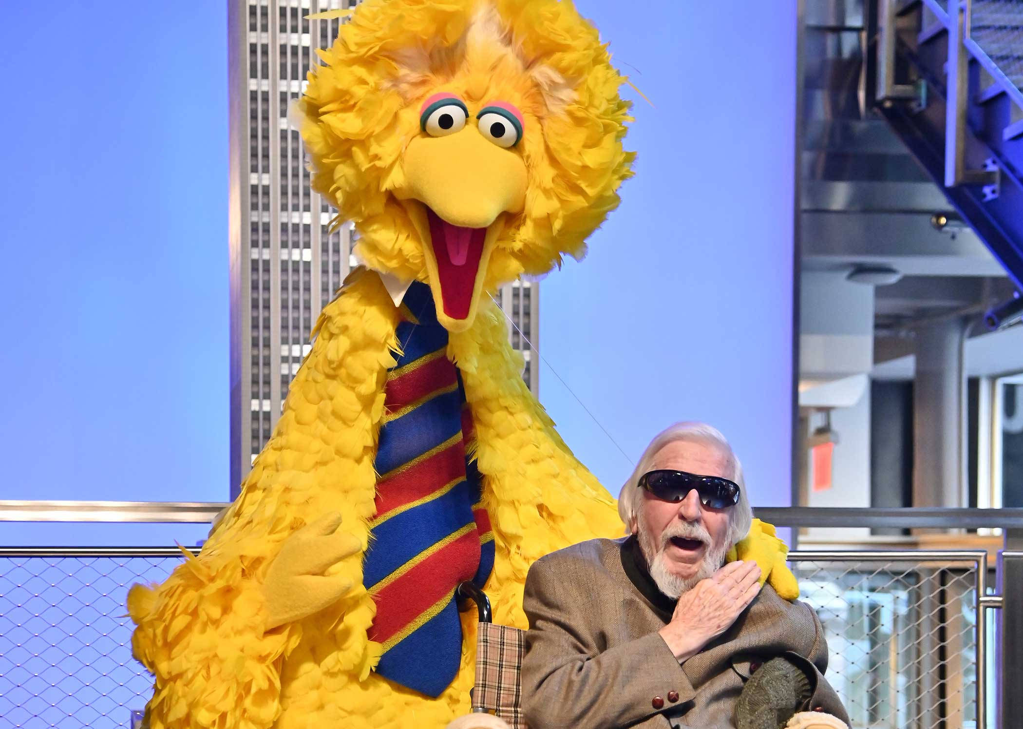 Big Bird 桌布 壁紙 — 一名戴太陽眼鏡的男子坐在巨型黃色大鳥布偶旁邊，布偶戴著條紋領帶，背景是城市。