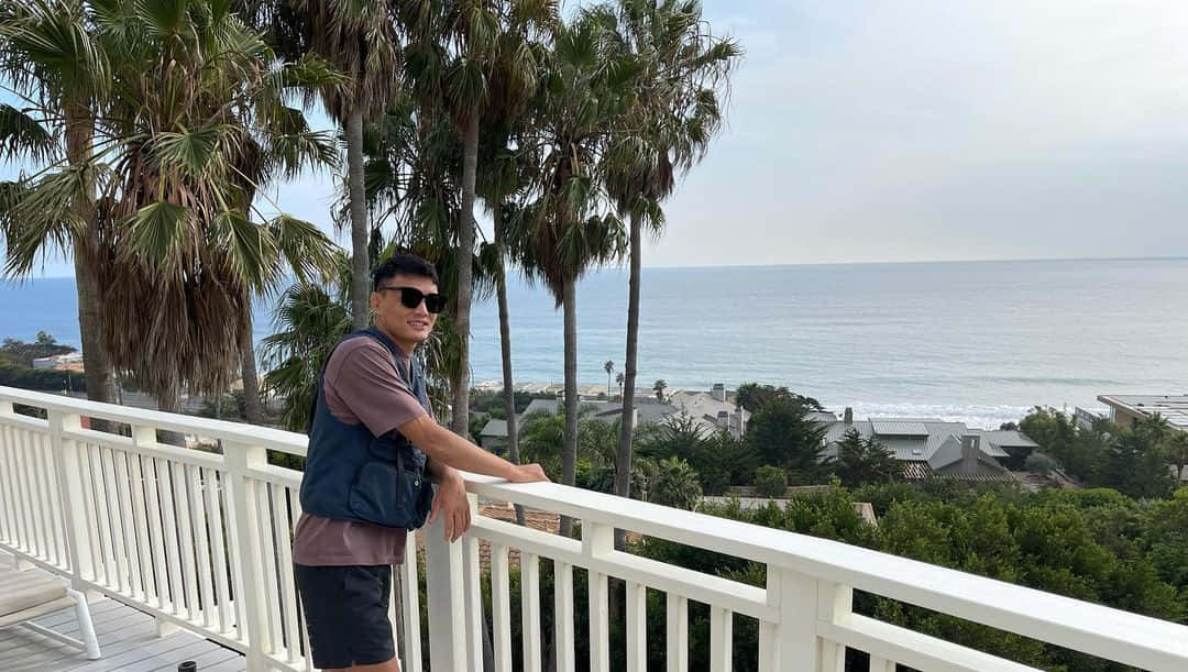 海灘 桌布 壁紙 — 一名戴太陽眼鏡的男子靠在白色陽台欄杆上，俯瞰下方平靜的海洋、棕櫚樹和沿海房屋。